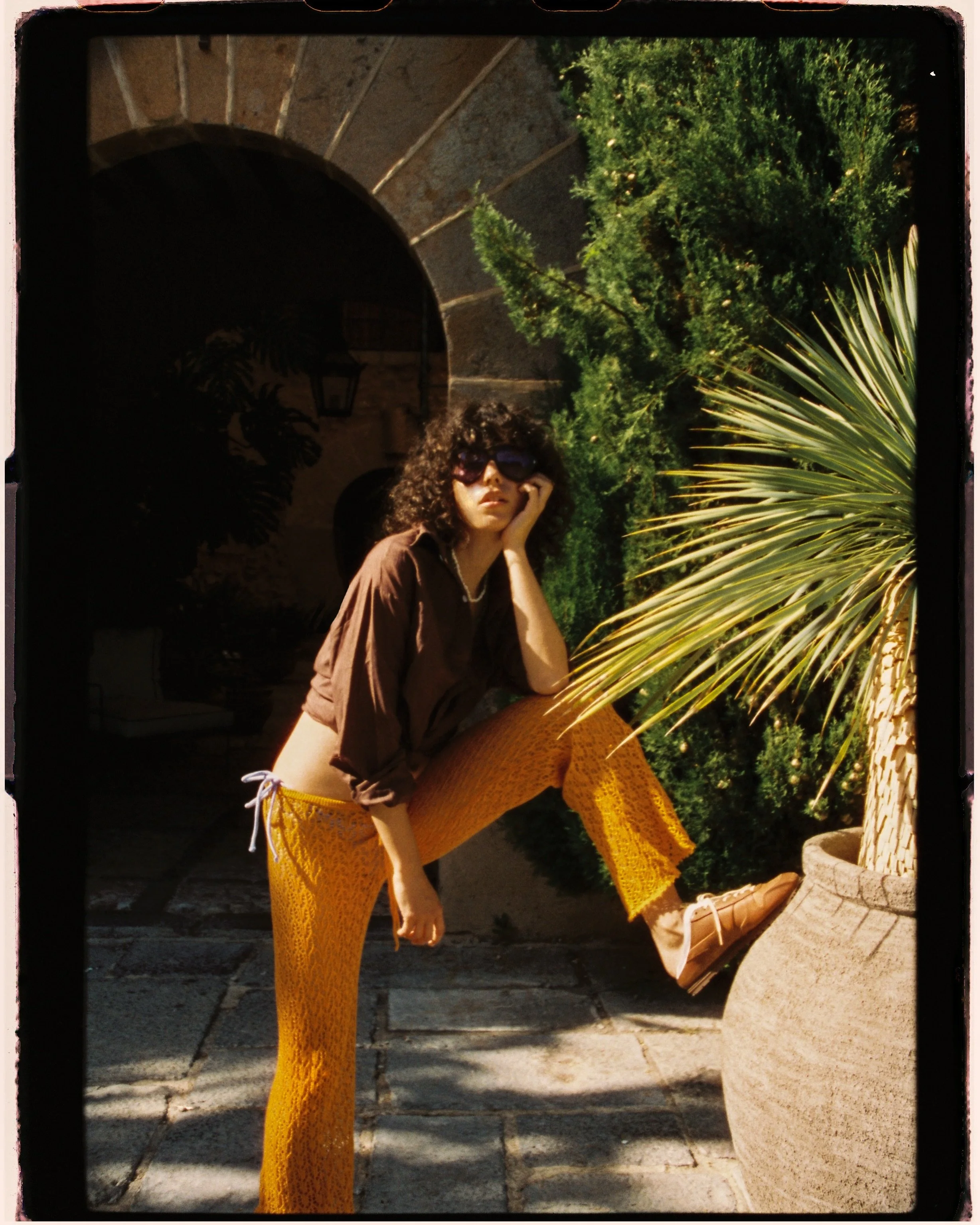 A woman with curly hair sitting outdoors, wearing sunglasses, a brown shirt, and yellow patterned pants, resting her head on her hand. She is surrounded by greenery, including a large potted plant and trees, underneath an arched stone structure.