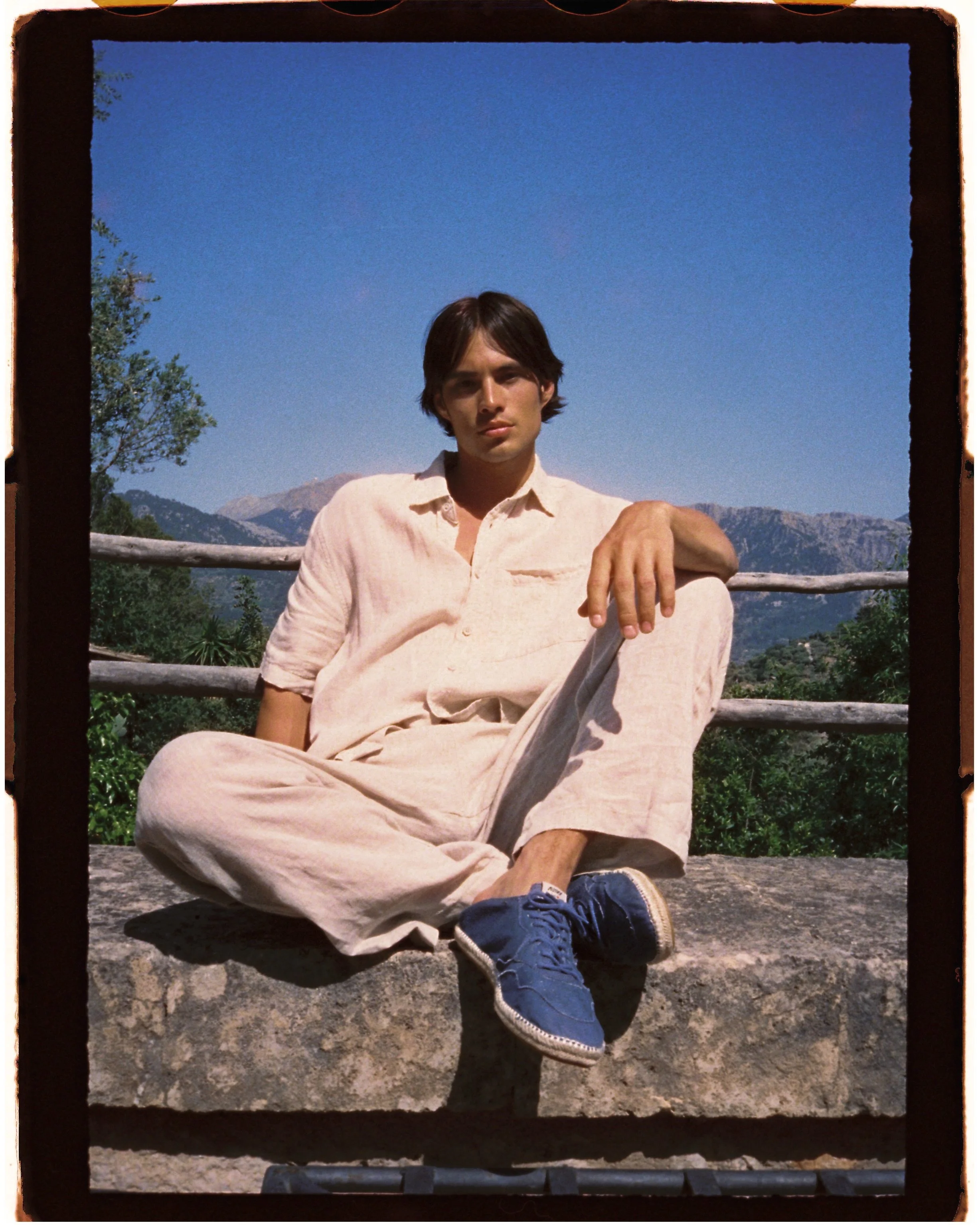 A young man with dark hair wearing a light-colored shirt and beige pants, sitting outdoors on a stone surface with grass and mountains in the background under a clear blue sky.