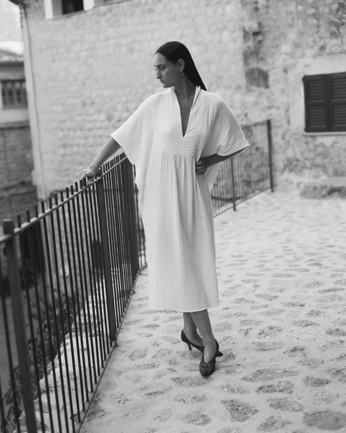 A woman with long dark hair, wearing a loose, light-colored dress and high heels, standing on a sandy path by a metal fence, gazing downward, with a stone building in the background.