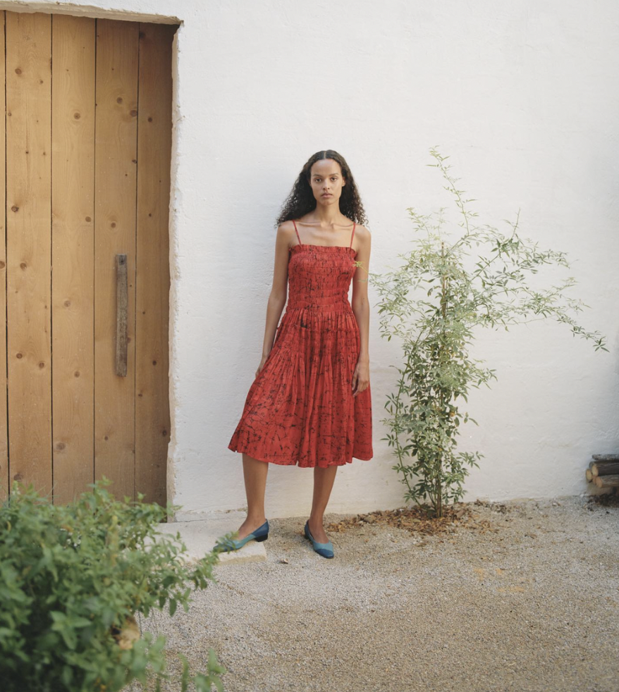 A woman in a red dress standing outdoors near a white wall, a wooden door, and some green plants.