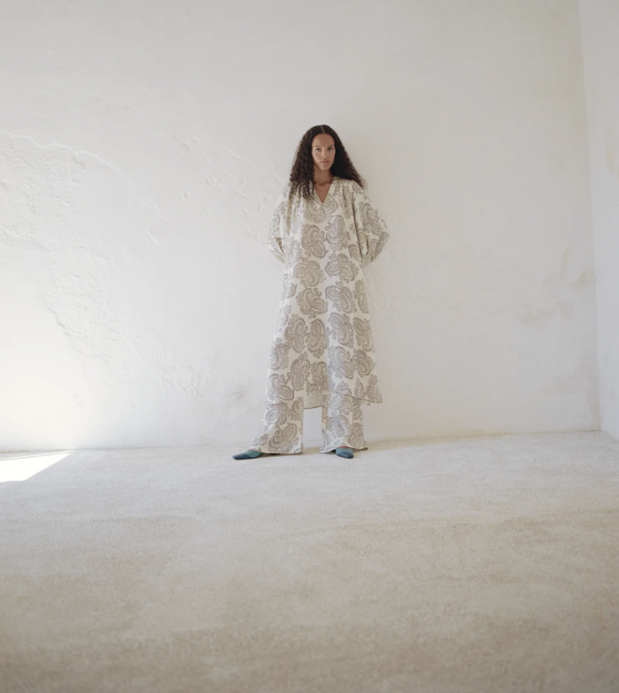 Woman standing against a textured white wall in a long, patterned beige and white dress in a minimalist setting with neutral tones.