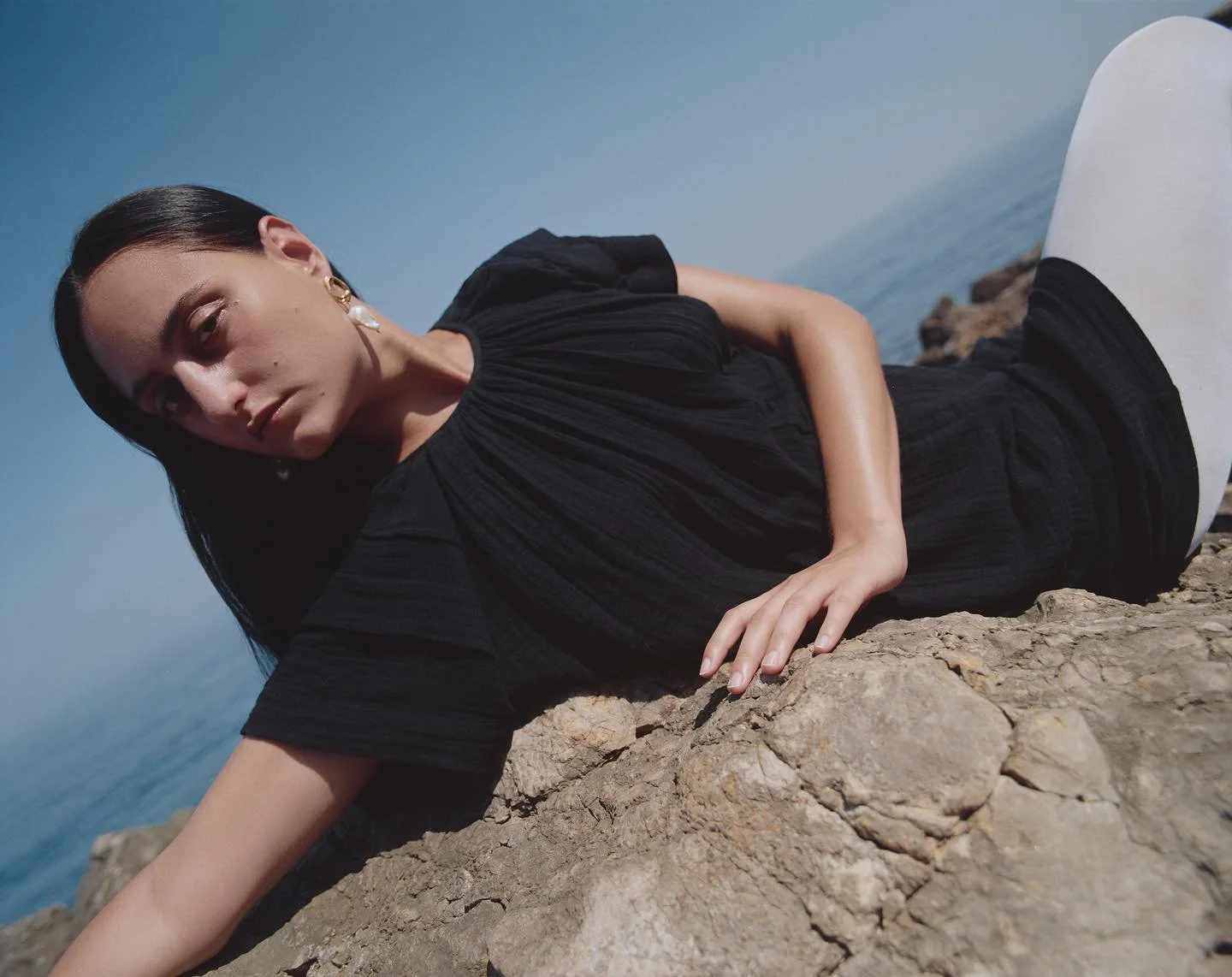 Woman in black dress lying on rocky surface near the ocean during daytime.