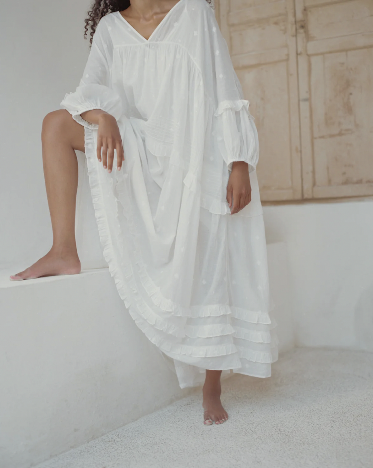A woman dressed in a flowing white dress with ruffled sleeves and layers, standing on a white surface in a minimalistic room with light-colored walls and a wooden cabinet.