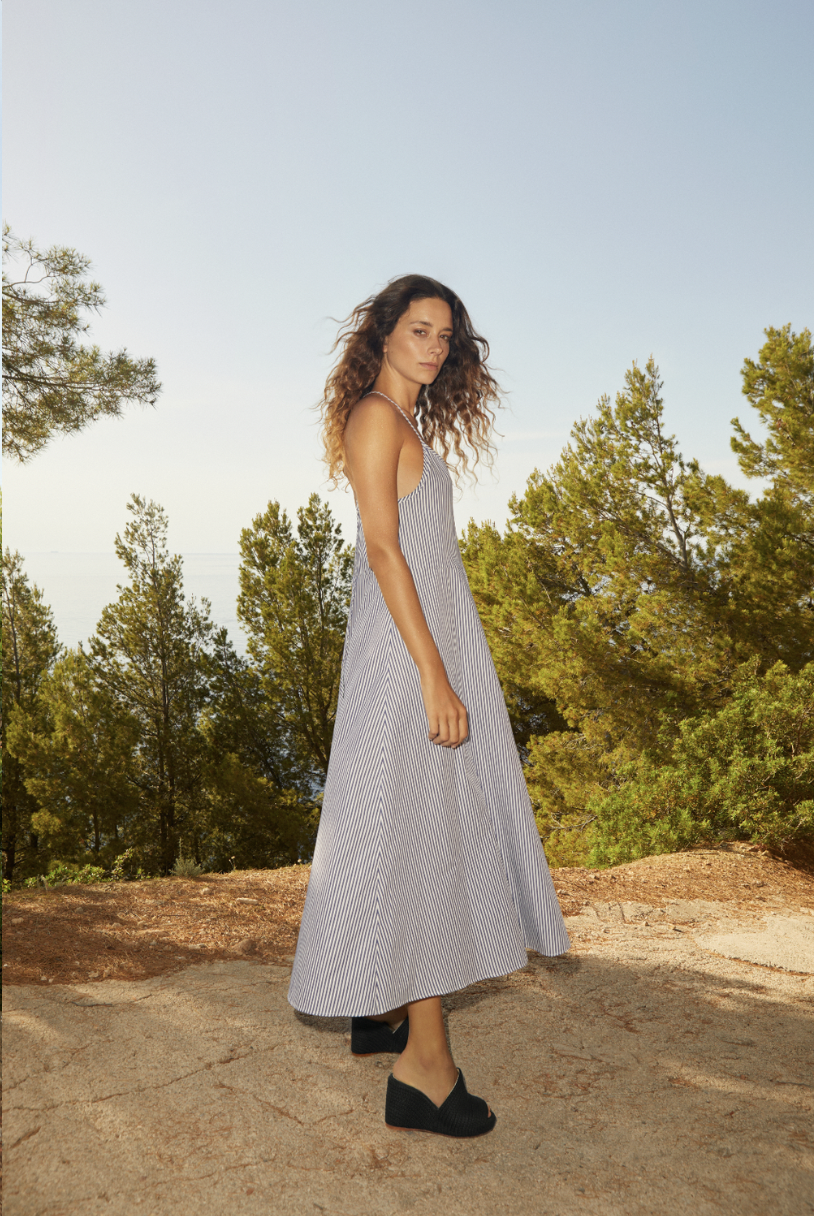 A woman with long curly hair wearing a sleeveless, striped, ankle-length dress and black wedge shoes standing outdoors on a rocky surface with trees and a clear sky in the background.
