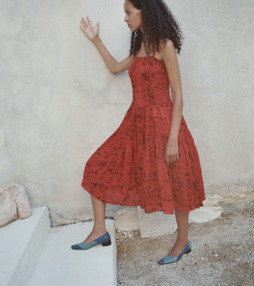 A woman with curly hair wearing a red textured sleeveless dress and blue shoes stands against a plain wall, with one foot on a white step and her hand touching the wall.