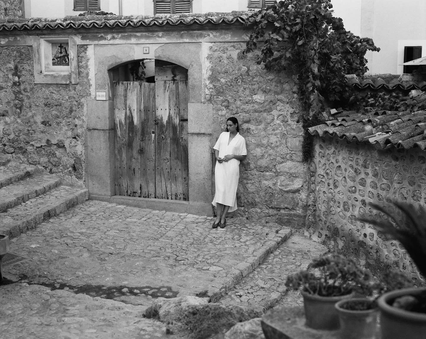 A woman in a white dress stands leaning against a stone wall near a wooden gate in a rustic setting, with cobblestone ground and tiled rooftops in the background.