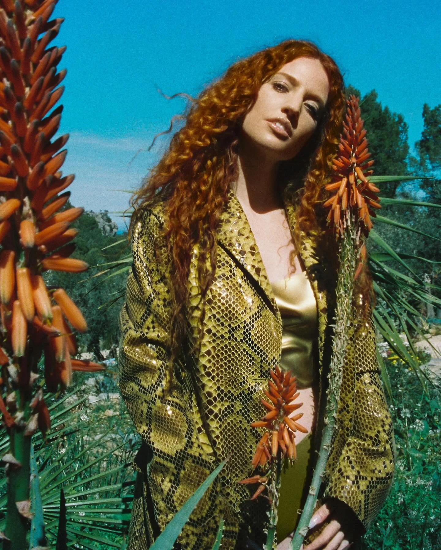 A woman with long curly red hair standing in a lush green outdoor area with plants and trees, wearing a shiny gold snakeskin-patterned jacket and a light-colored top.