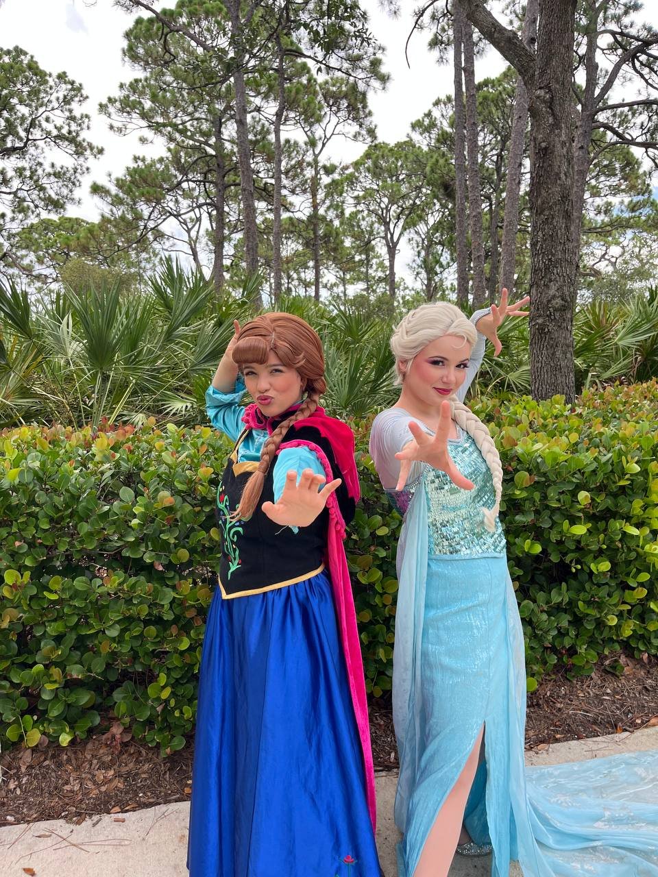 Two women dressed as princesses, one as Anna from Frozen and the other as Elsa from Frozen, striking playful poses outdoors in front of bushes and trees.