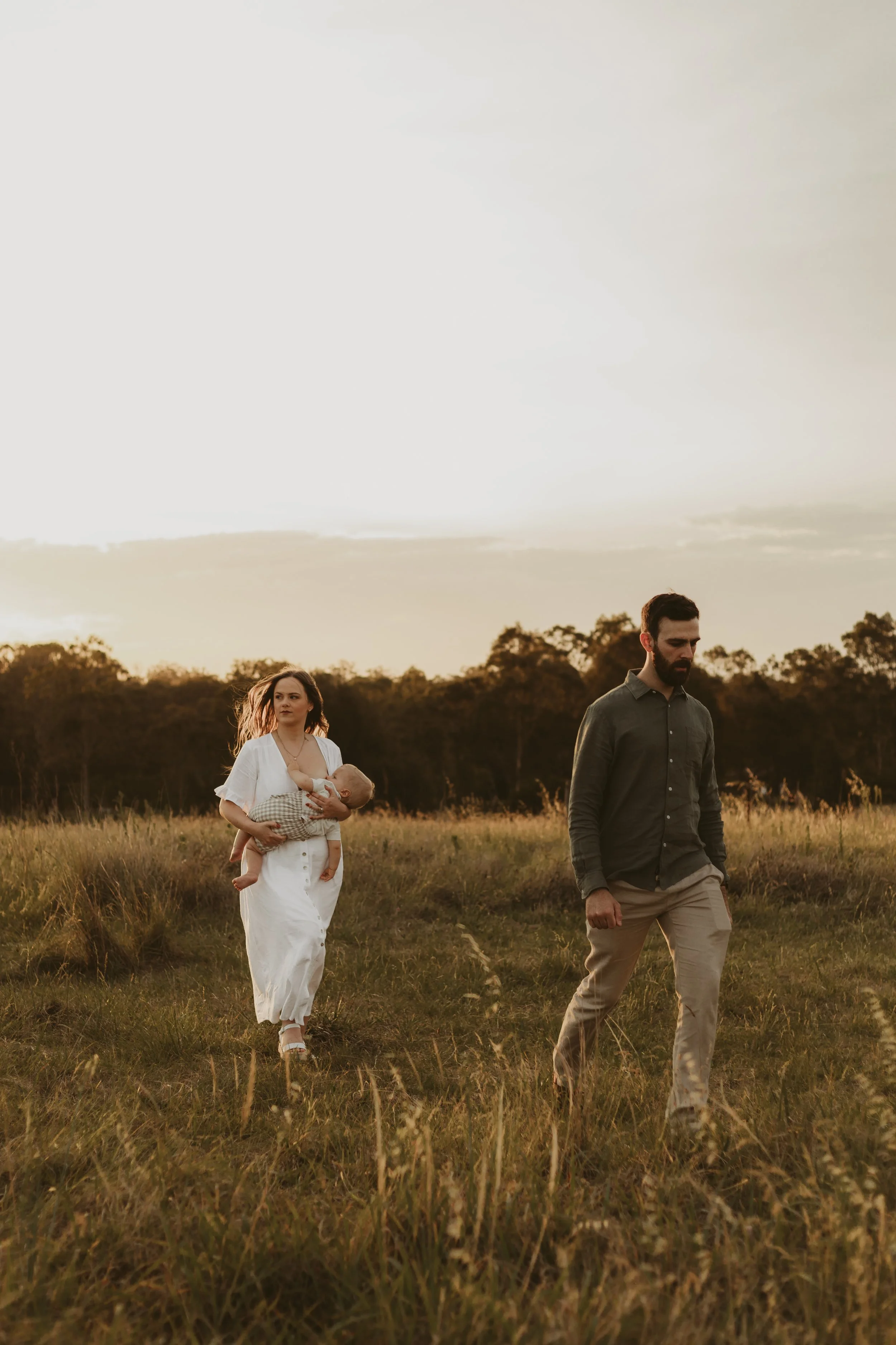 A woman in a white dress carrying a baby in a checkered outfit, walking in a grassy field with a man in a dark shirt and beige pants nearby, during sunset with trees in the background.