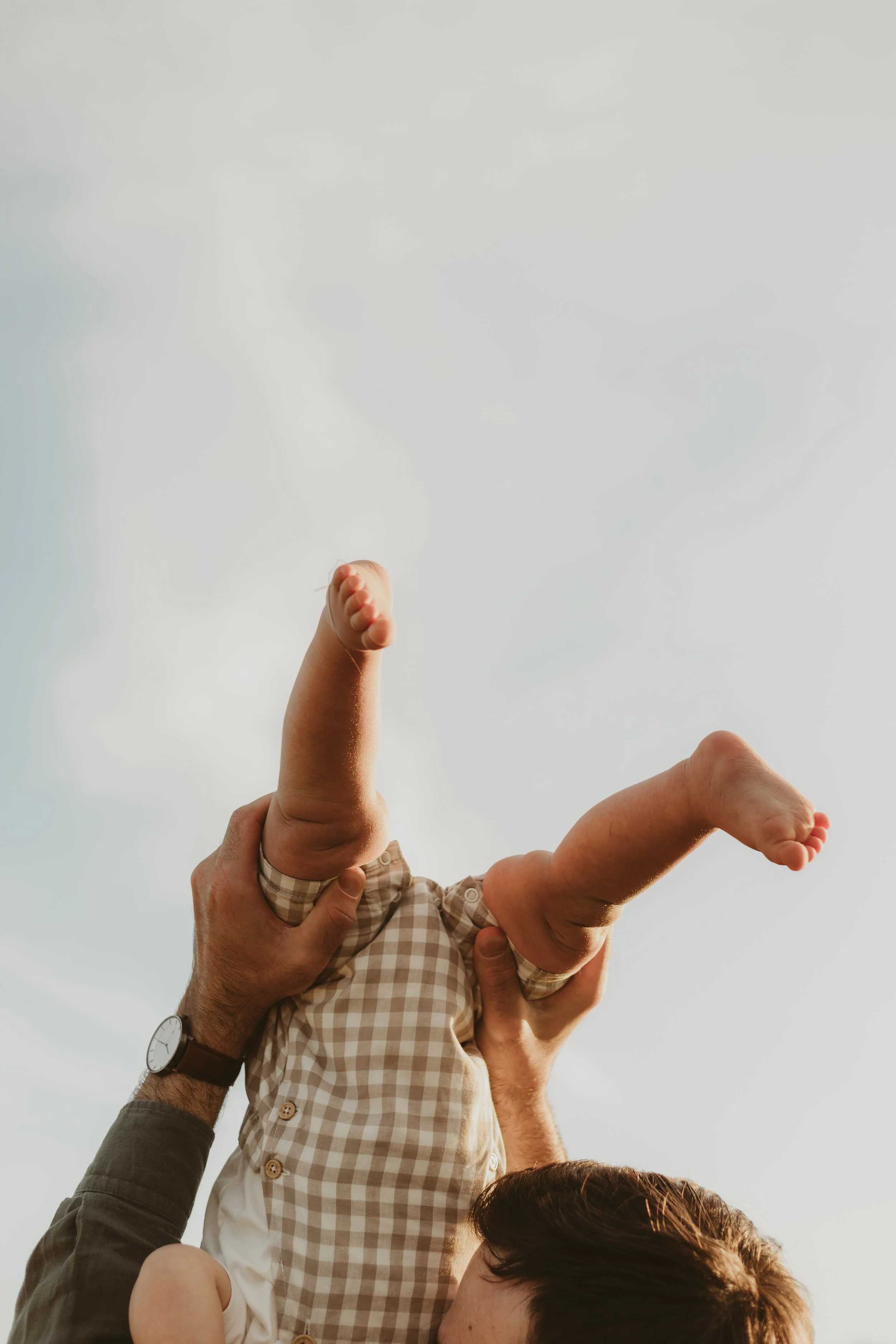 Adult holding a baby upside down outdoors against a cloudy sky