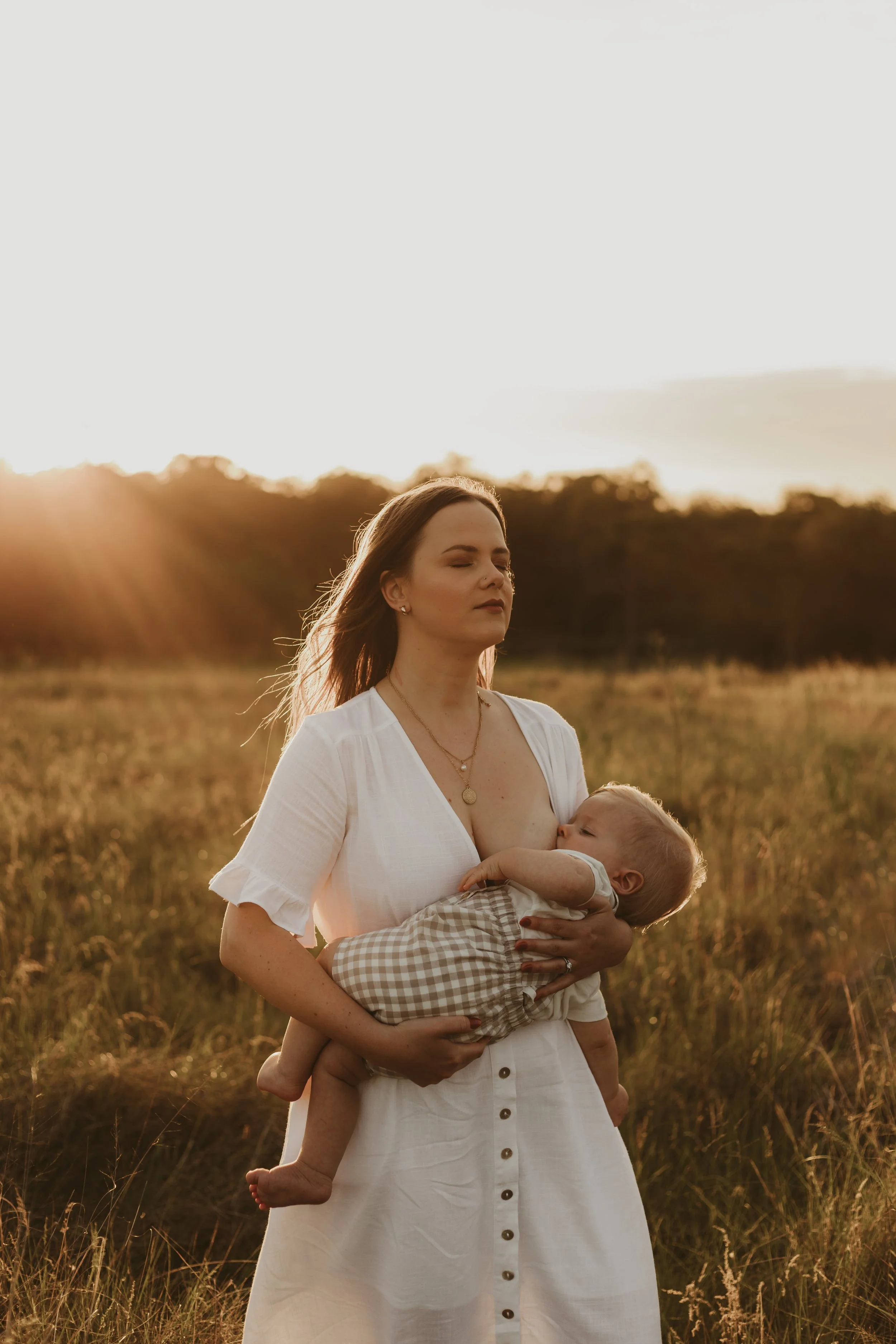 A woman holding a young child in a grassy field during sunset, with warm light and a soft-focus background of trees.