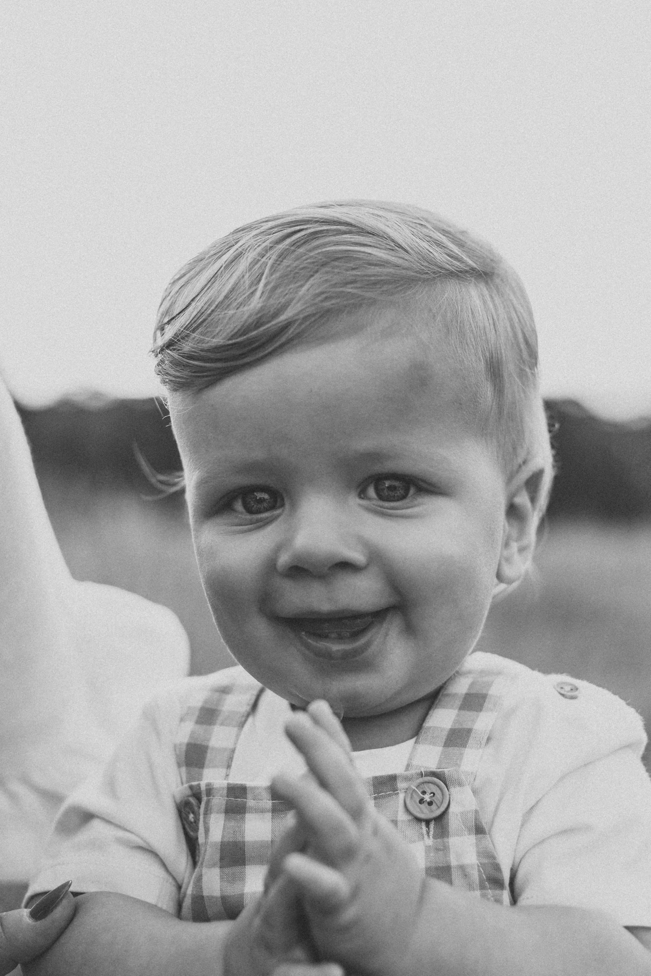 Black and white photo of a young smiling boy with short hair, holding his hands together in front of him, outdoors with blurred background.