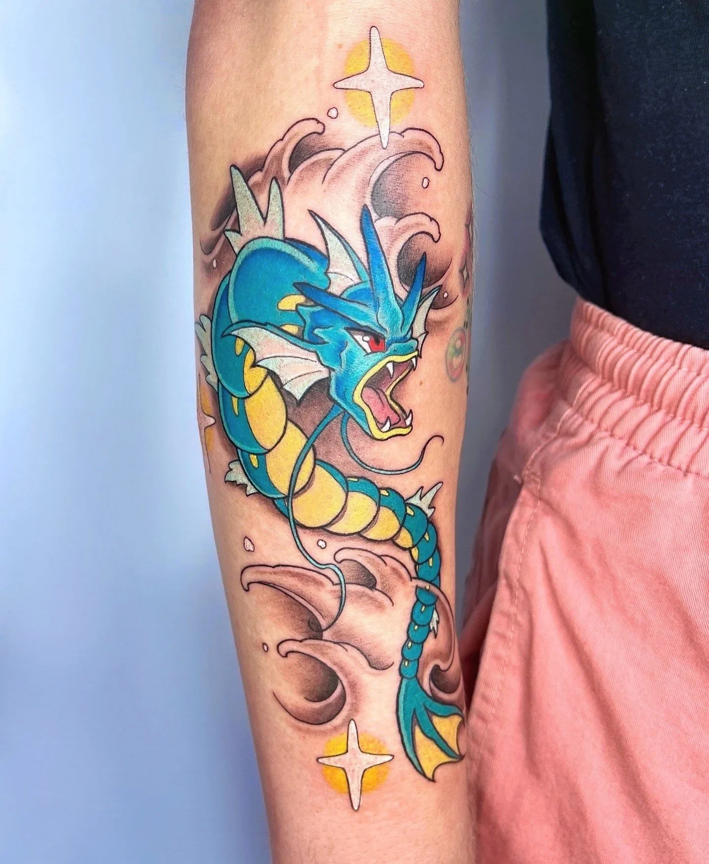 Colorful tattoo of a blue and yellow dragon with an open mouth showing sharp teeth, set on the person's upper arm, with clouds, steam, and star-like symbols around it.