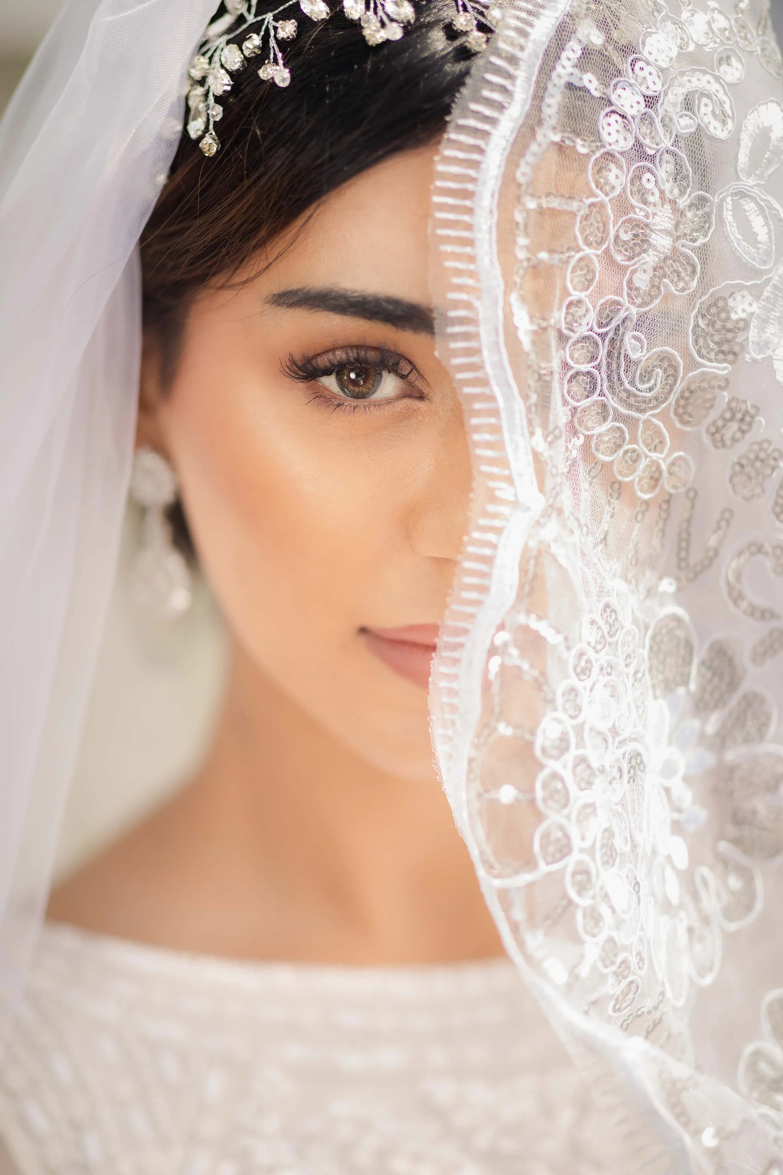 Close-up of a bride's face, partially covered with an ornate lace veil featuring embroidery and sequins, with her brown eye and dark eyebrow visible.