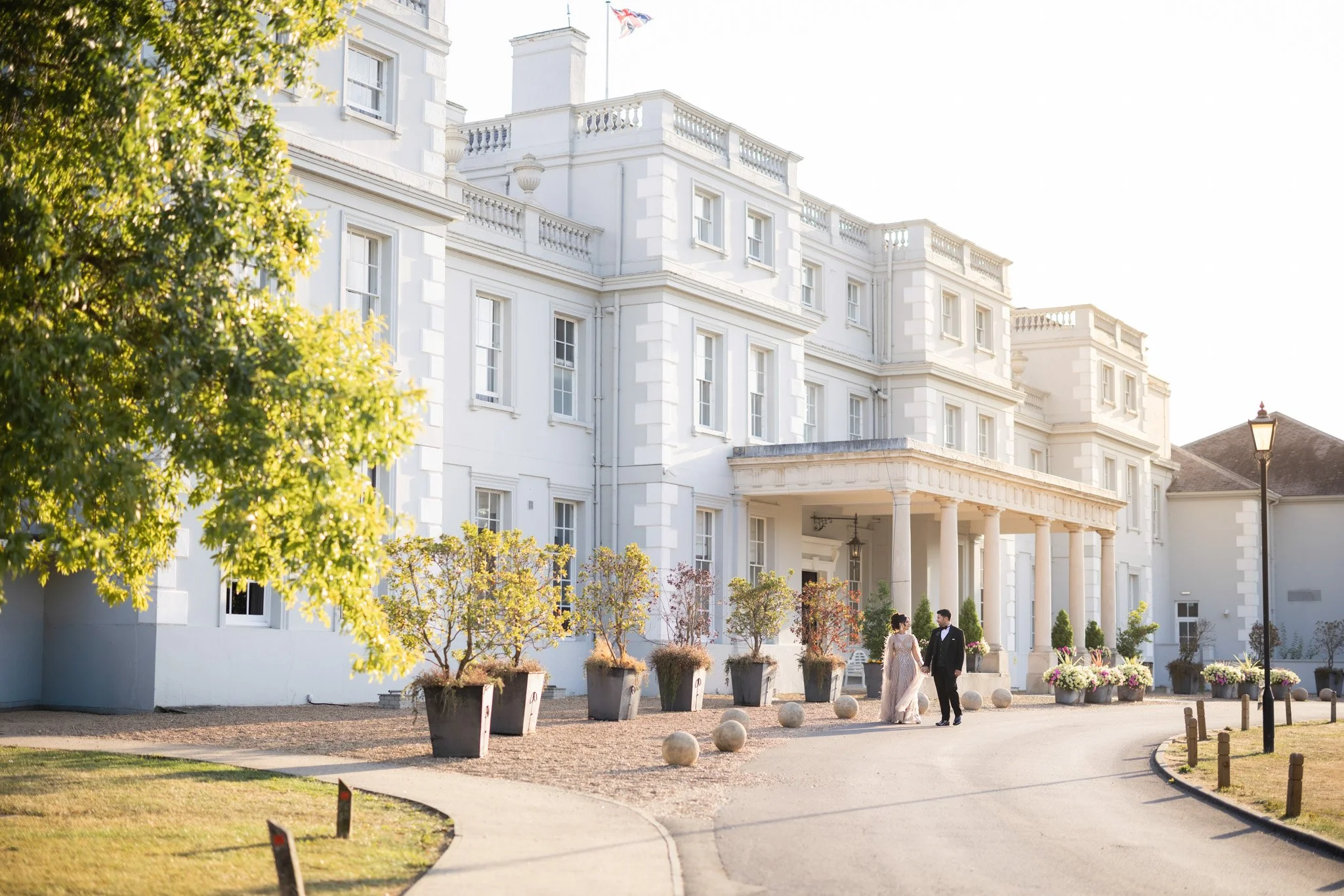 A white mansion with multiple stories and columns at the entrance. Two people in formal attire are walking and talking in front of the mansion, surrounded by potted plants and decorative spheres on the ground. The setting appears to be during the daytime with a clear sky.