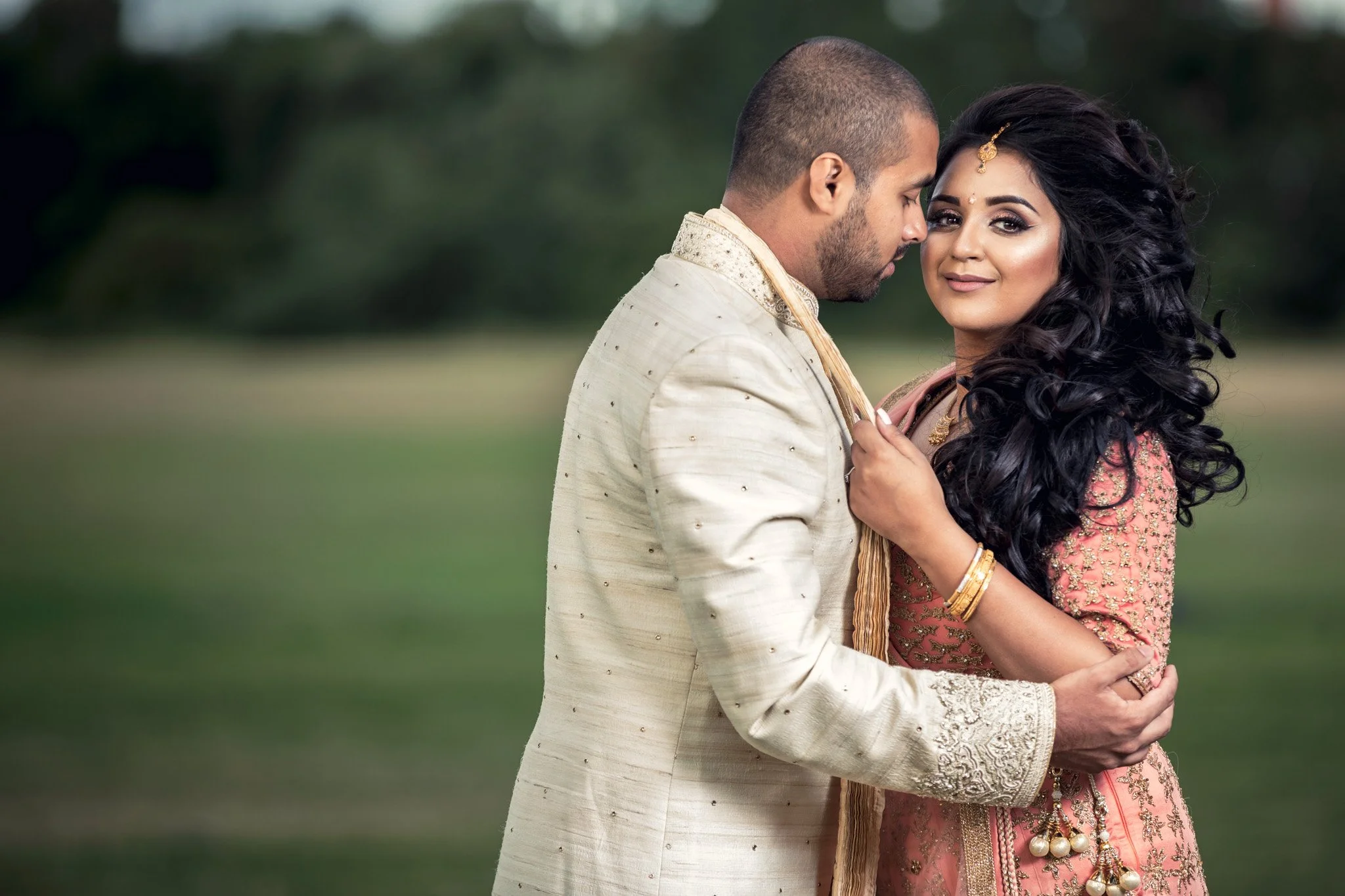 A couple dressed in traditional Indian wedding attire embracing outdoors with a blurred natural background.