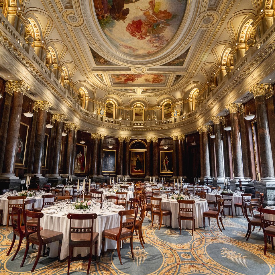 Elegant banquet hall with round tables covered in white tablecloths, set with glassware and candles, surrounded by wooden chairs. The hall features dark marble columns with gold accents, a high ornate ceiling with fresco artwork, and framed paintings on the walls.
