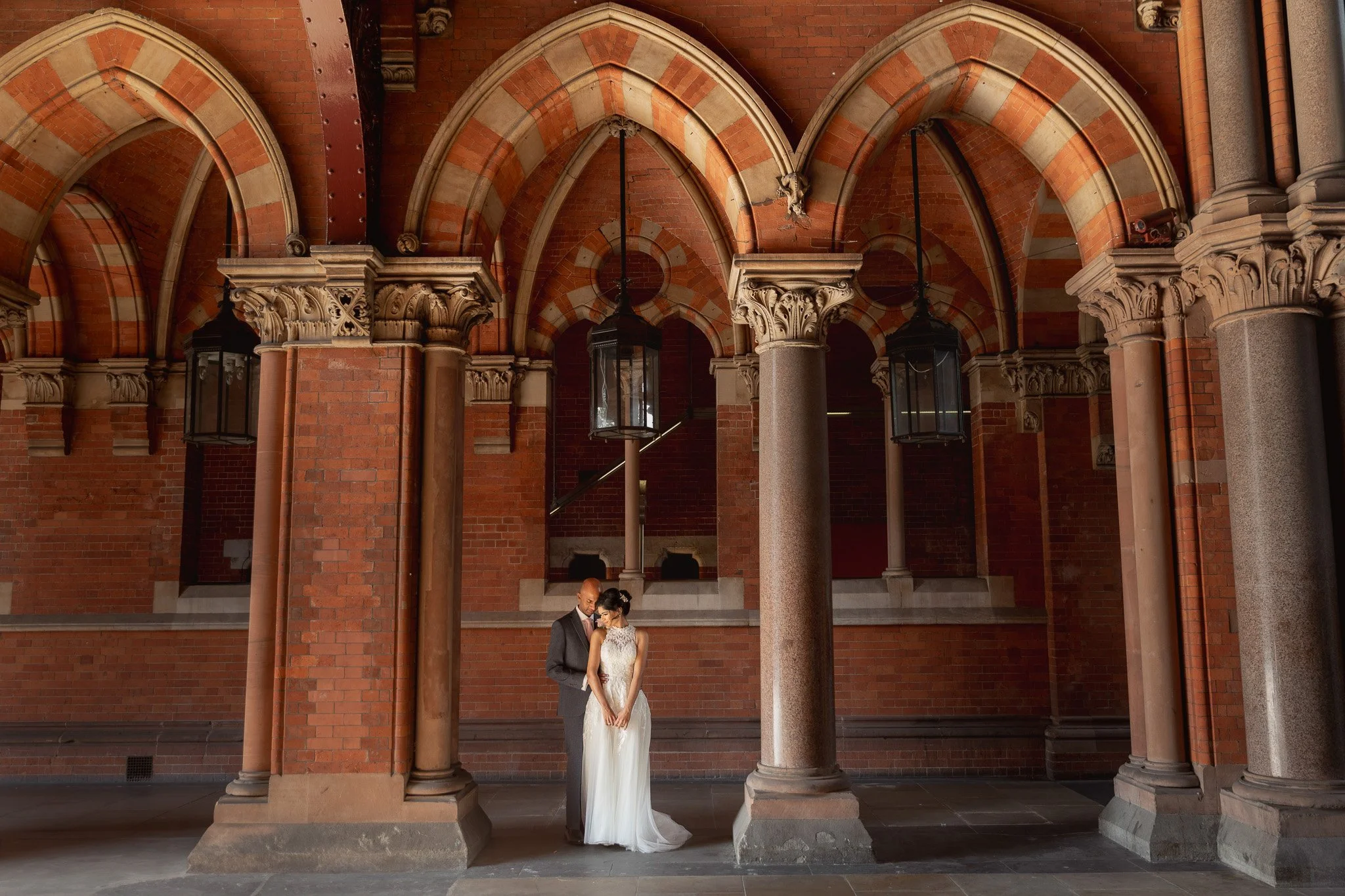 A bride and groom standing close together under gothic arches made of red brick and stone columns at night.