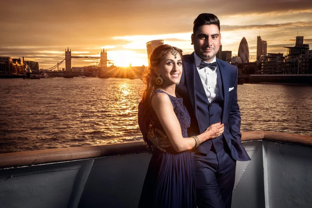 A couple dressed in formal attire standing on a boat with London skyline and Tower Bridge at sunset in the background.