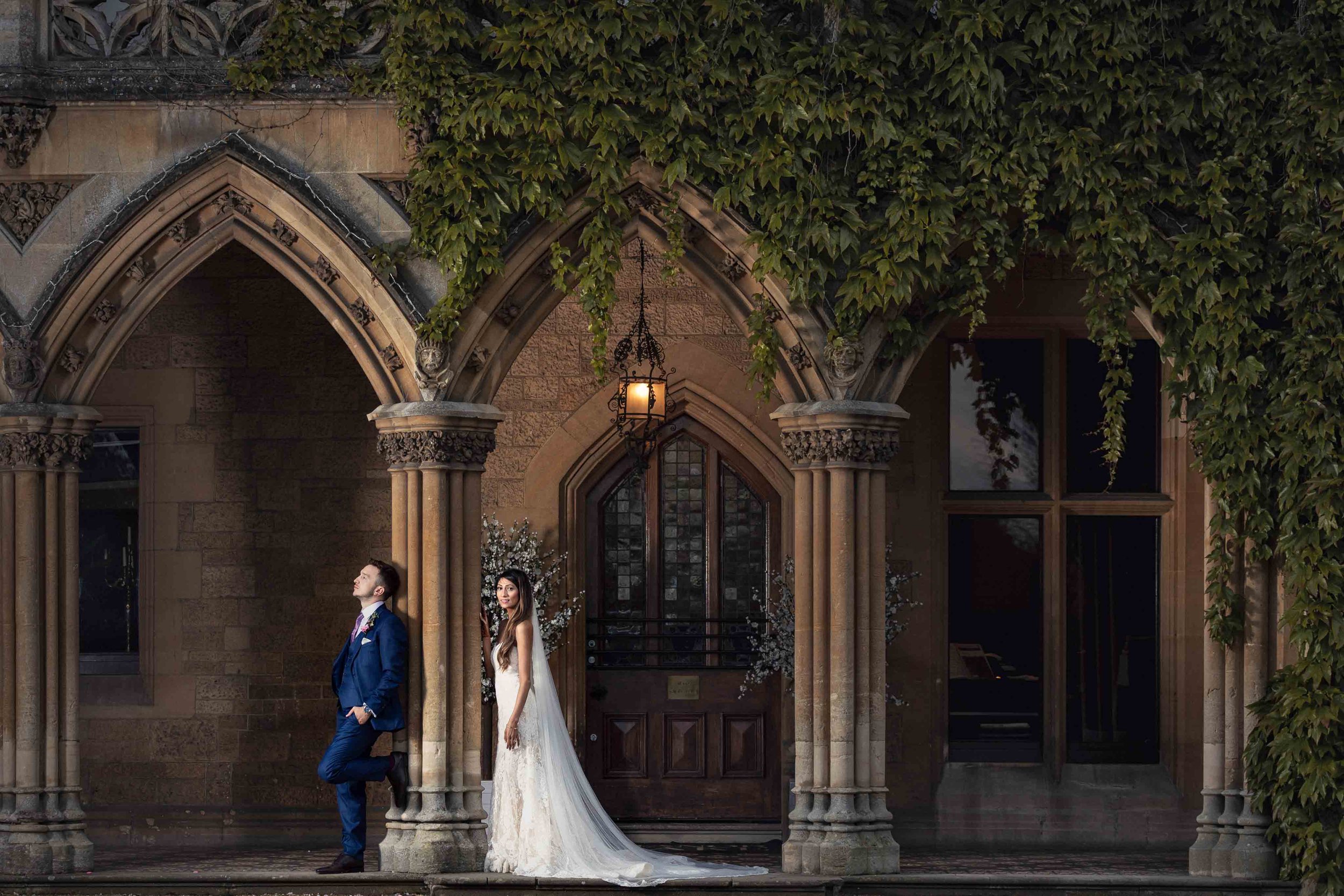 A bride in a white wedding dress and a groom in a blue suit stand outside a Gothic-style stone building with arches and ivy, at dusk.