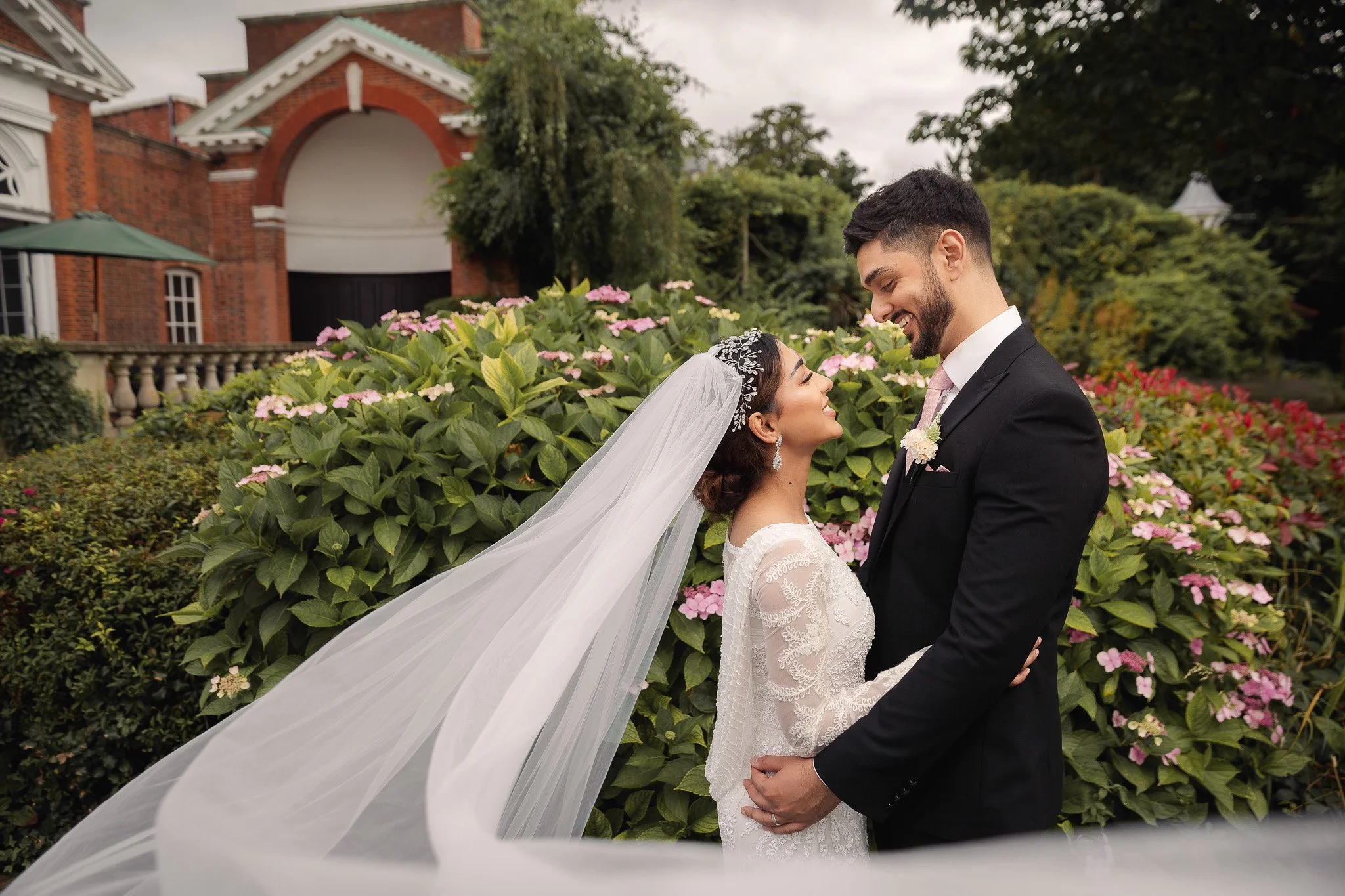 A bride and groom standing close together outdoors, smiling at each other, with a lush garden and a brick building in the background.