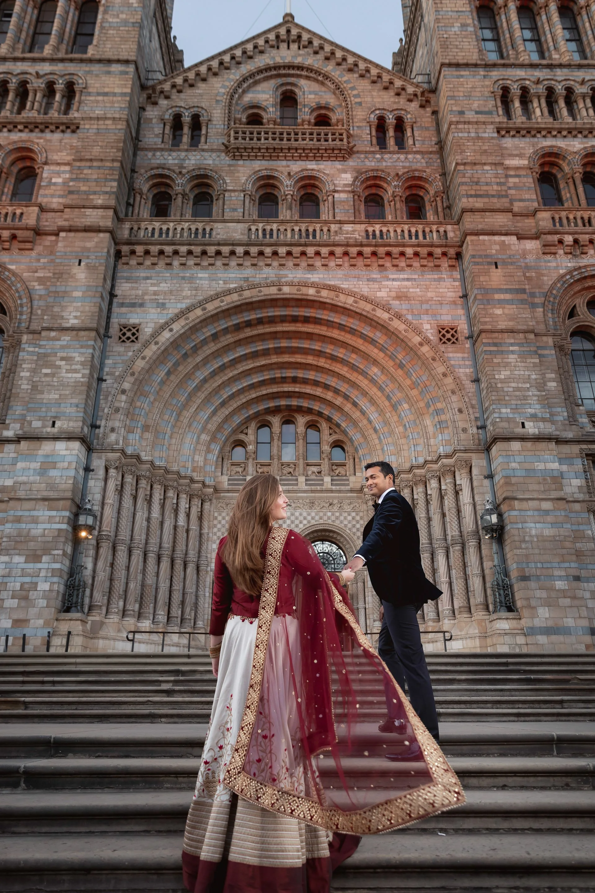 A couple dressed in formal attire, with the woman in traditional Indian clothing and the man in a suit, holding hands and standing on the steps in front of a historic, ornate brick building with arched entrance, during sunset.
