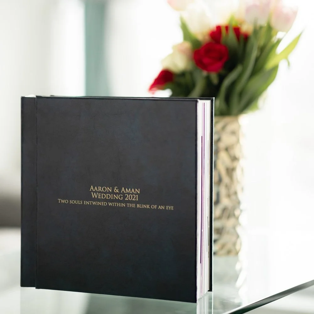 Wedding photo album with a black cover, titled "Aaron & Aman Wedding 2021; Two Souls Entwined Within the Blink of an Eye," placed on a reflective surface with a blurred bouquet of flowers in the background.