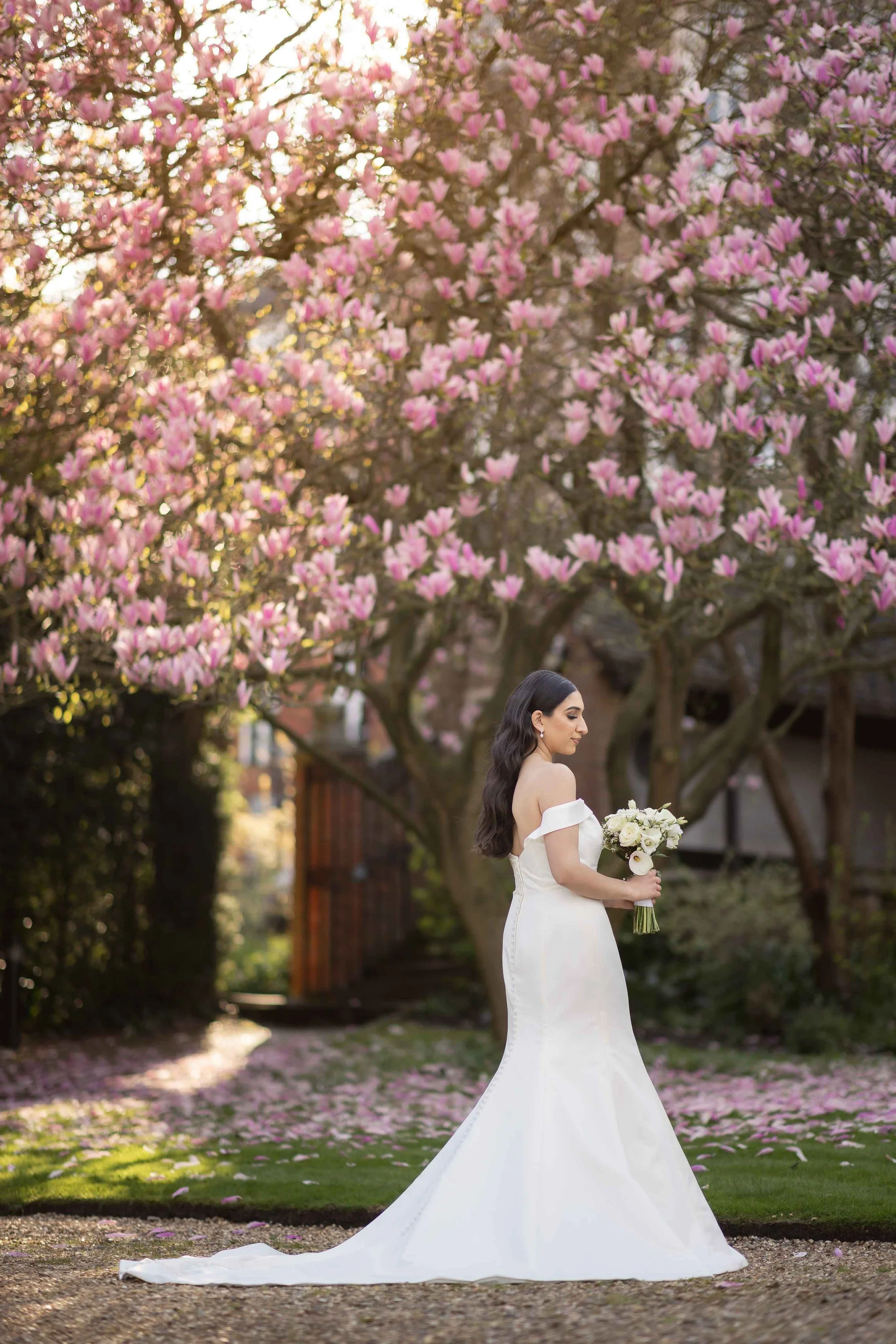 A bride in a white wedding dress holding a bouquet of white flowers, standing outdoors under a blooming pink magnolia tree on a sunny day.