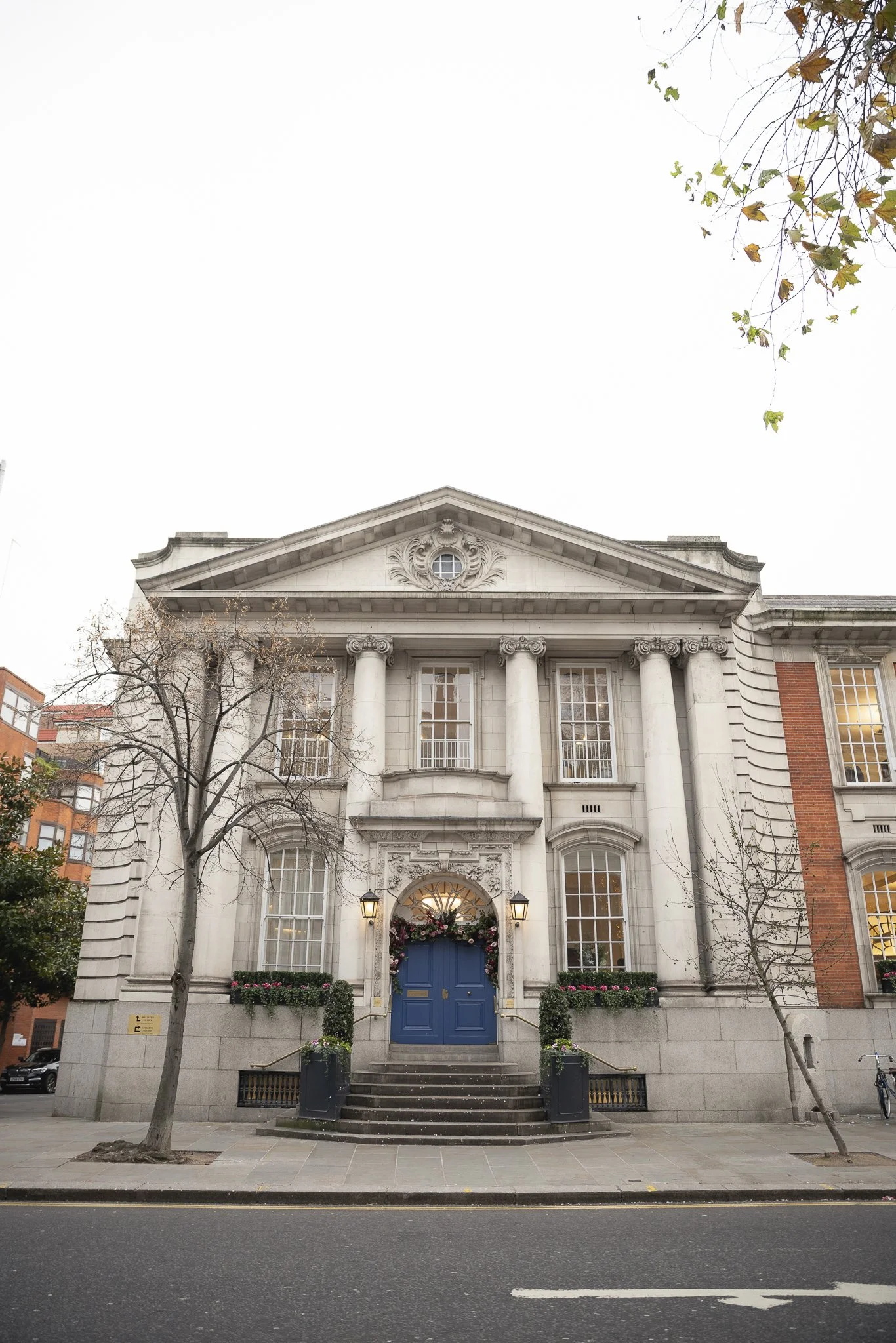 Front view of a grand, historic building with white columns, ornate detailing, and a blue door, decorated with holiday wreaths and flowers, located on a city street with a few leafless trees.
