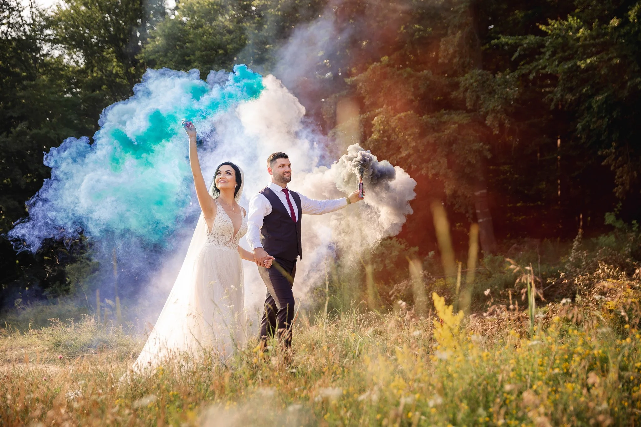A bride and groom outdoors holding smoke bombs that release colorful smoke, with a trail of blue, green, white, black, and yellow smoke, surrounded by trees and grass.