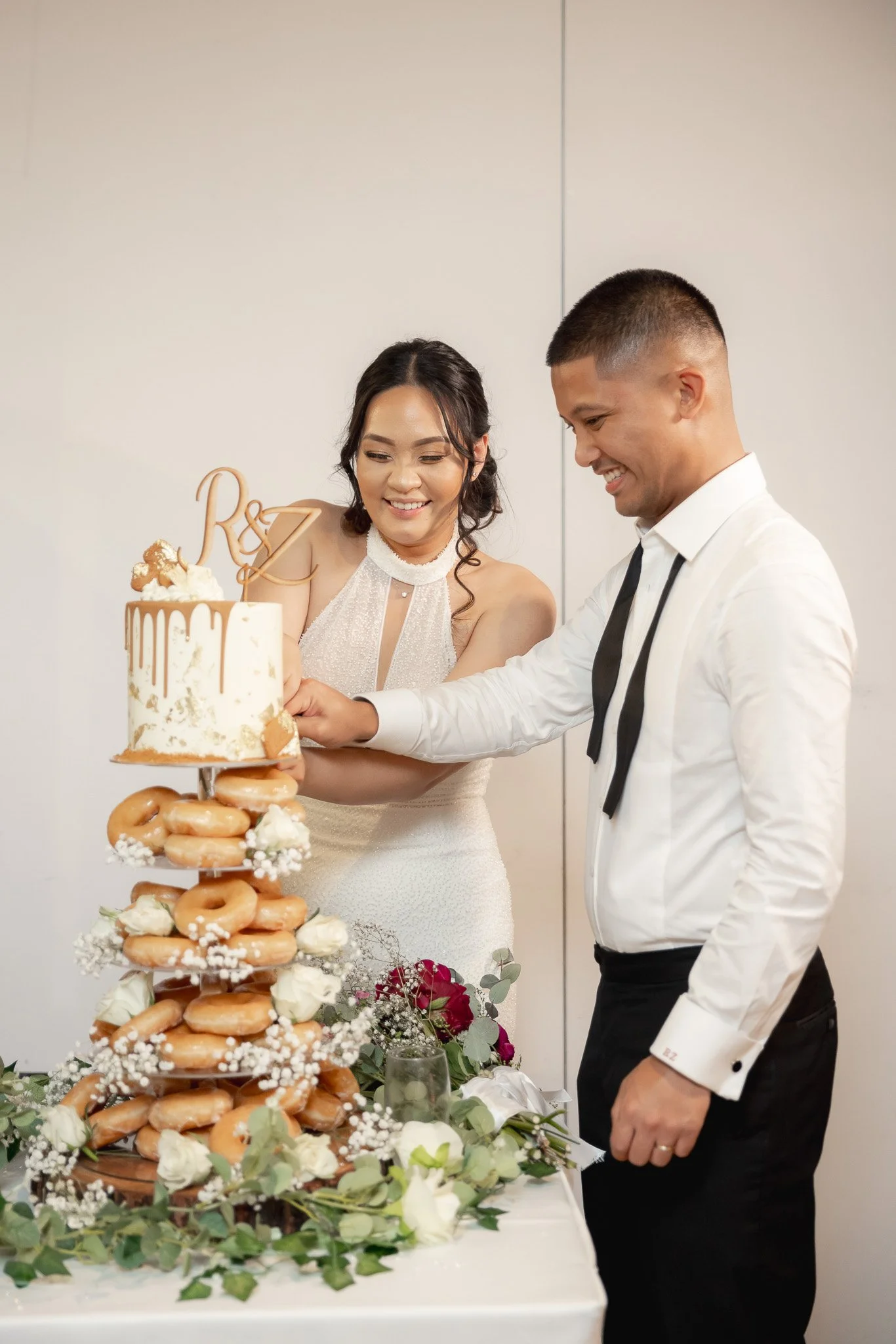 A couple cut a wedding cake at their reception. The cake is decorated with a topper featuring initials and is surrounded by donuts and flowers.