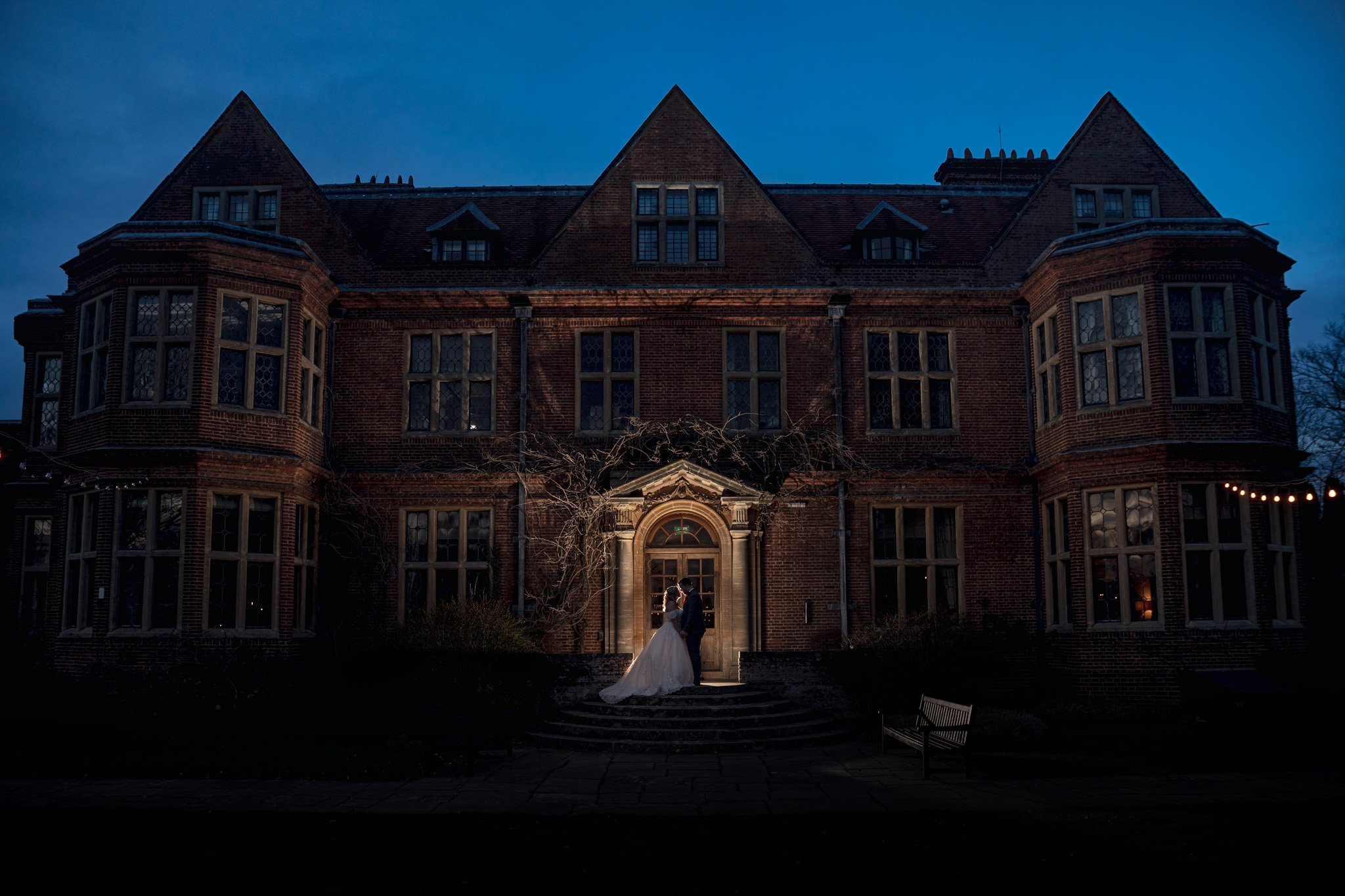 A bride and groom standing on steps in front of a large, old brick mansion at dusk, with string lights hanging on the right side.