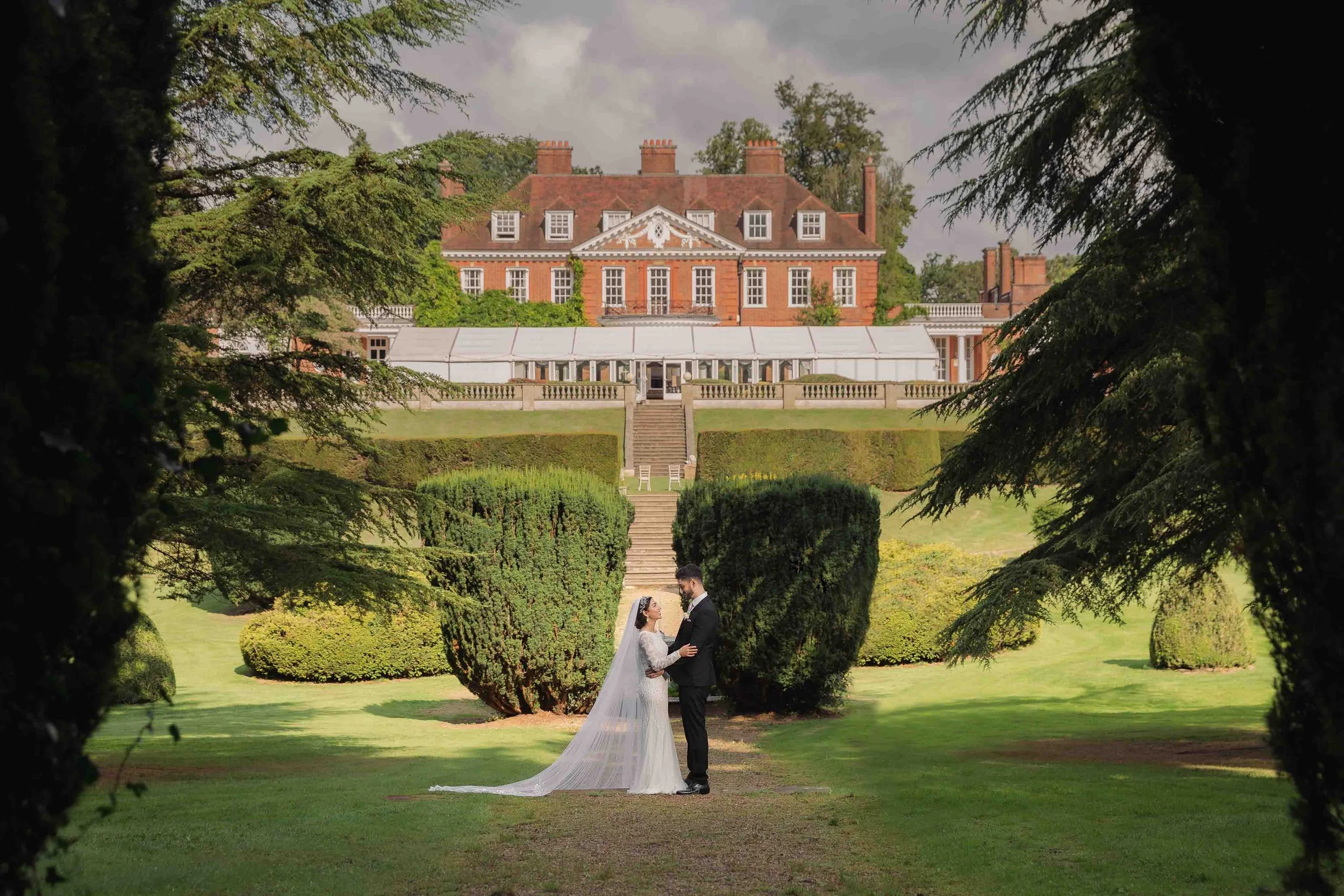 A bride and groom standing in a garden, holding hands and looking at each other, with a large historic mansion in the background. The bride is wearing a white wedding dress with a long veil, and the groom is in a black tuxedo. The garden is lush with