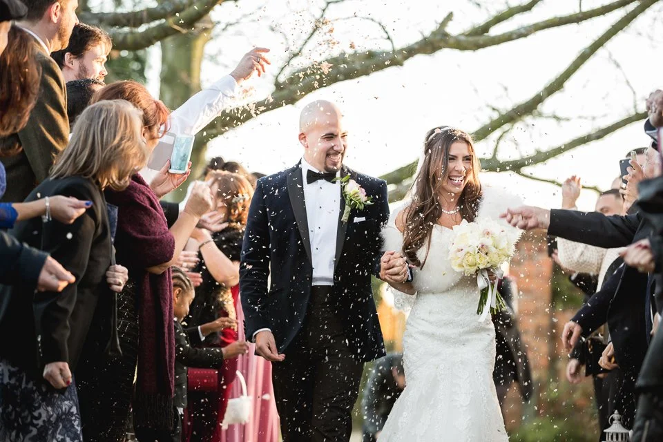 Bride and groom walking outdoors through a crowd of wedding guests who are throwing confetti or rice in celebration.