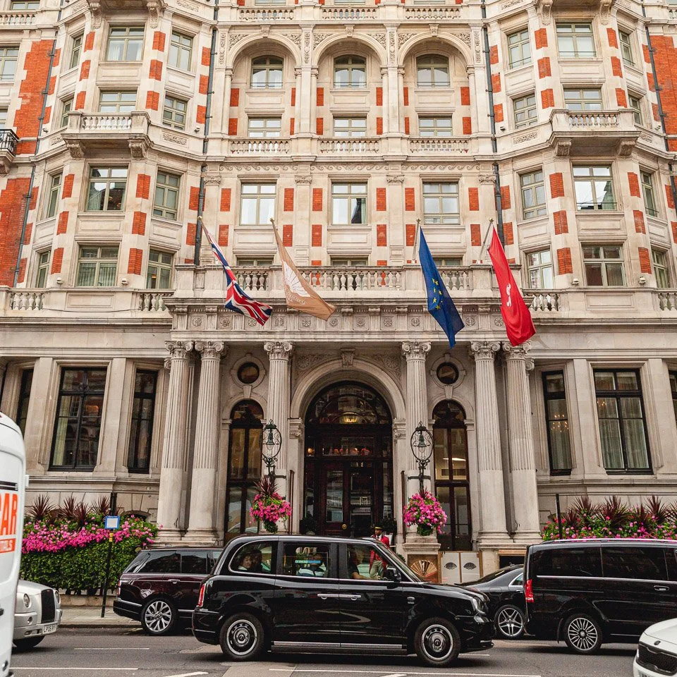 A grand, historic hotel building with ornate architectural details, multiple windows, and flags of the UK, EU, and local area hanging above the entrance. There are cars parked and driving on the street in front, with pink flowers and greenery on the sidewalk.