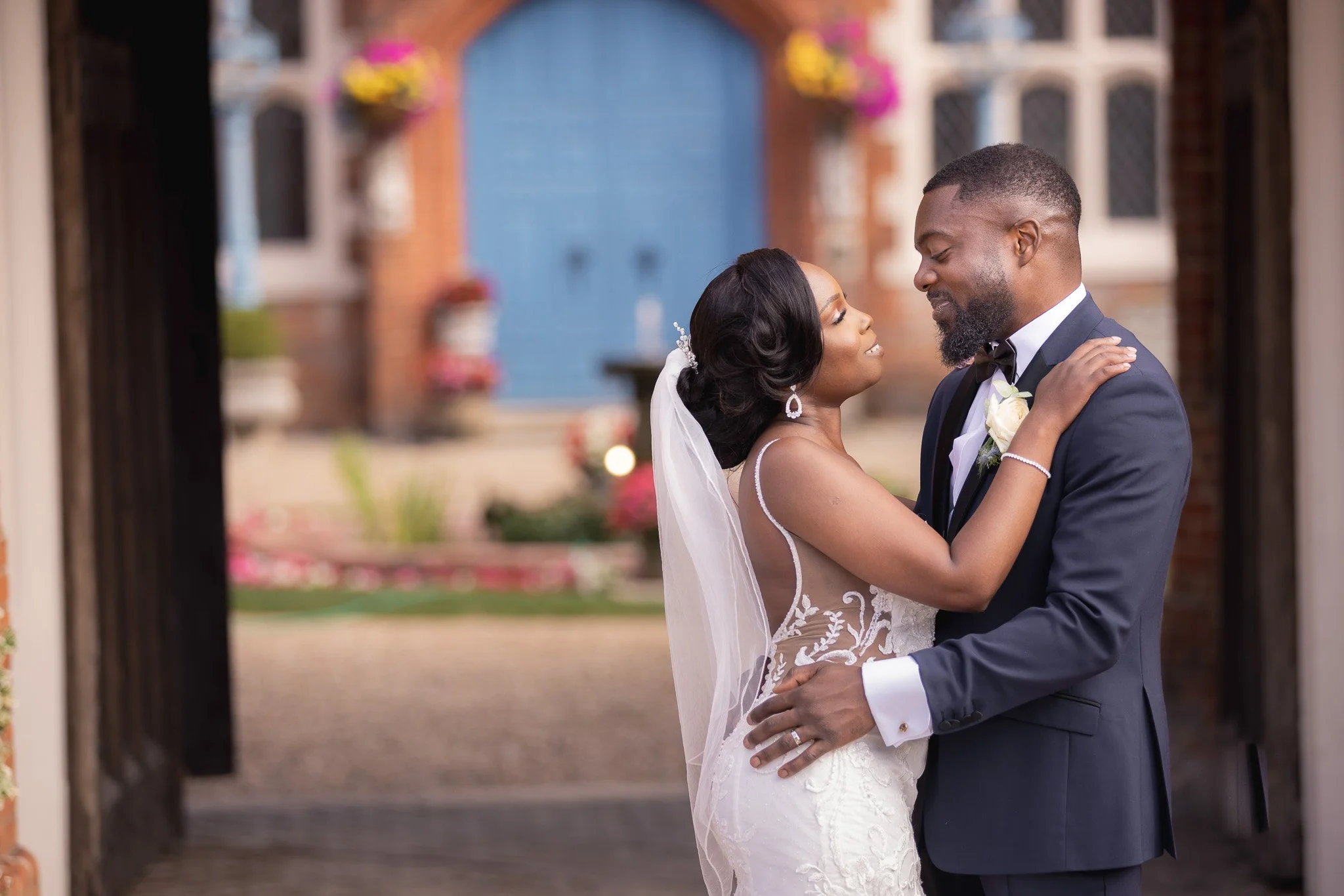 A bride and groom sharing a dance outside a building, with the bride wearing a lace wedding gown and the groom in a tuxedo, looking at each other lovingly.