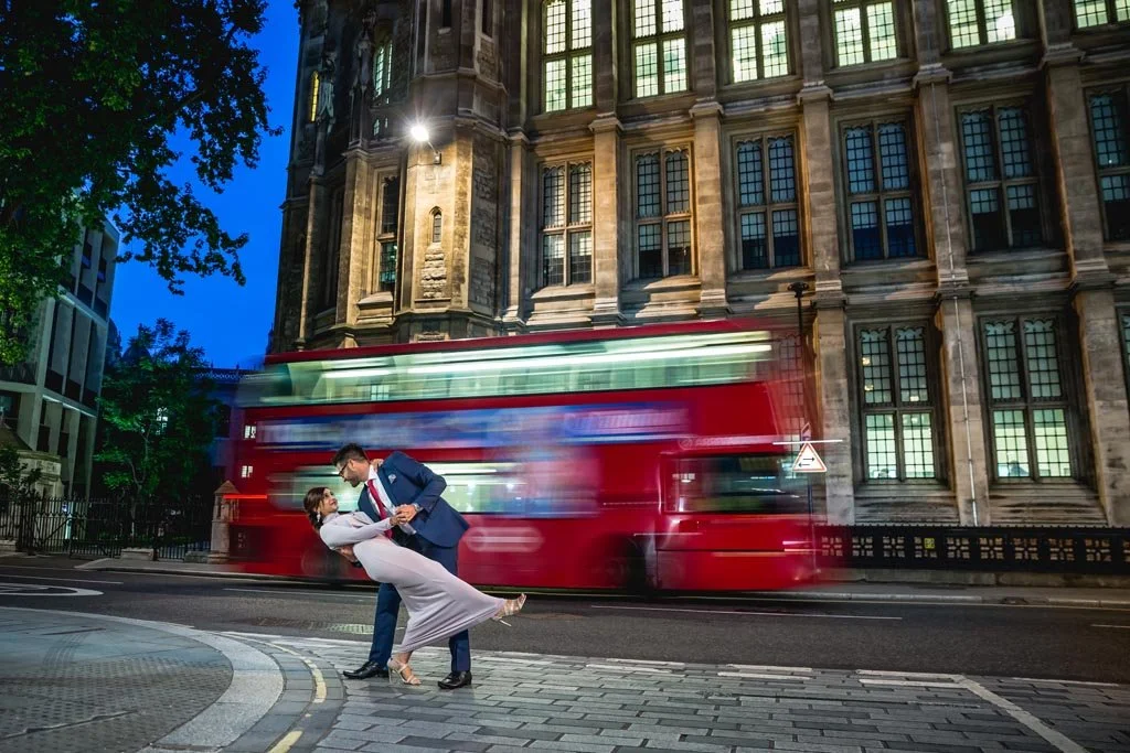 A man in a suit dips a woman in a dress while dancing on a city street at dusk, with a blurred red double-decker bus passing by in the background and a historic building with large windows behind them.