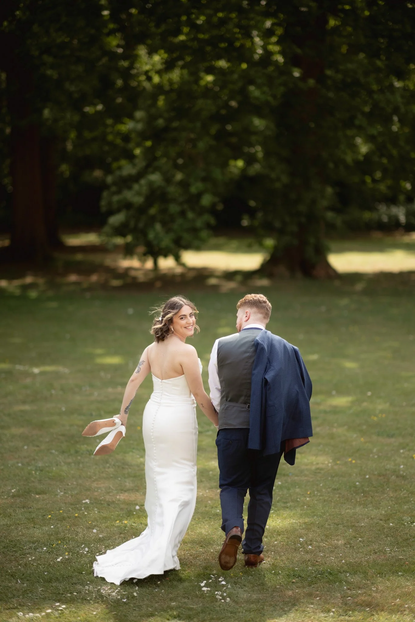 A couple in wedding attire walking hand in hand in a park, with the woman smiling and looking back at the camera, holding her shoes in her left hand, and the man carrying a jacket over his shoulder.
