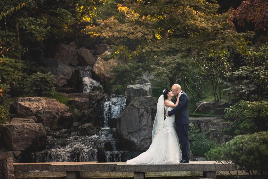 A bride and groom standing on a wooden platform embracing in front of a waterfall surrounded by lush greenery and trees.
