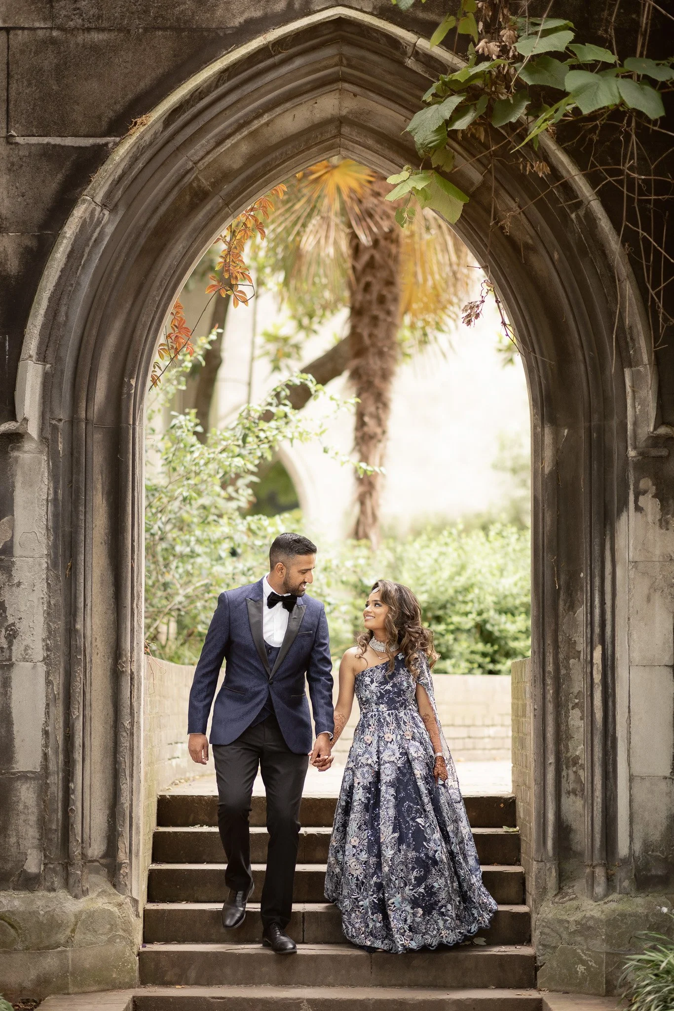 A couple dressed in formal attire, the man in a tuxedo and the woman in an elegant gown, holding hands and walking down stone steps through an arched stone gateway surrounded by lush greenery.