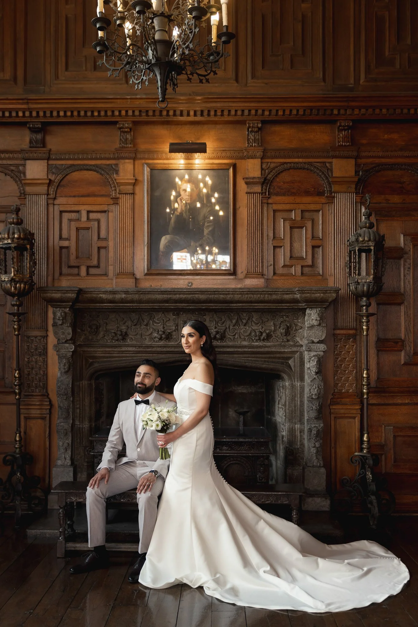 Bride and groom in formal wedding attire sitting in front of an ornate fireplace in a wood-paneled room, with a large portrait reflection of the photographer hanging above them.