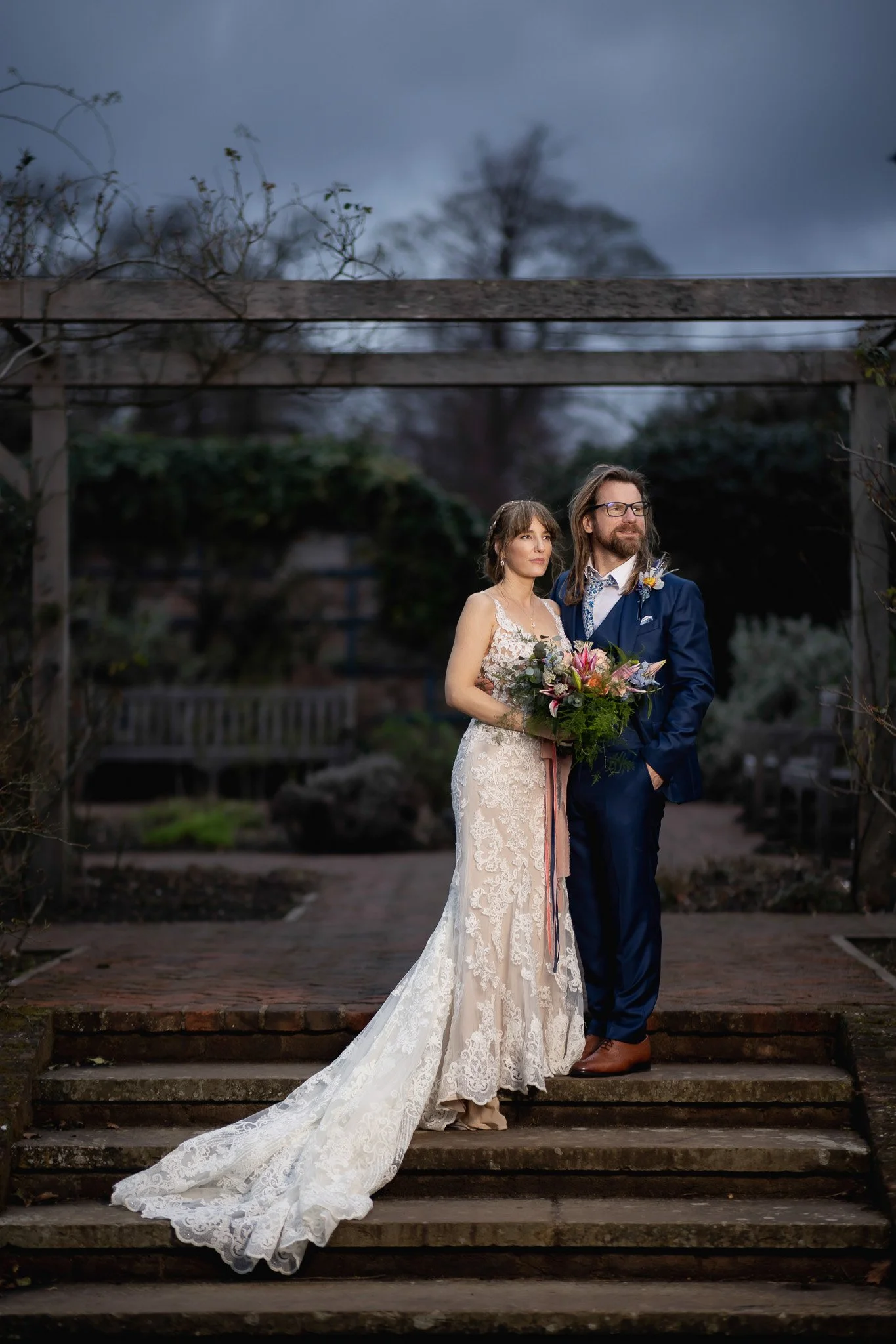 A bride in a lace wedding gown holding a bouquet, standing next to a groom in a blue suit with glasses, outdoors during cloudy weather.