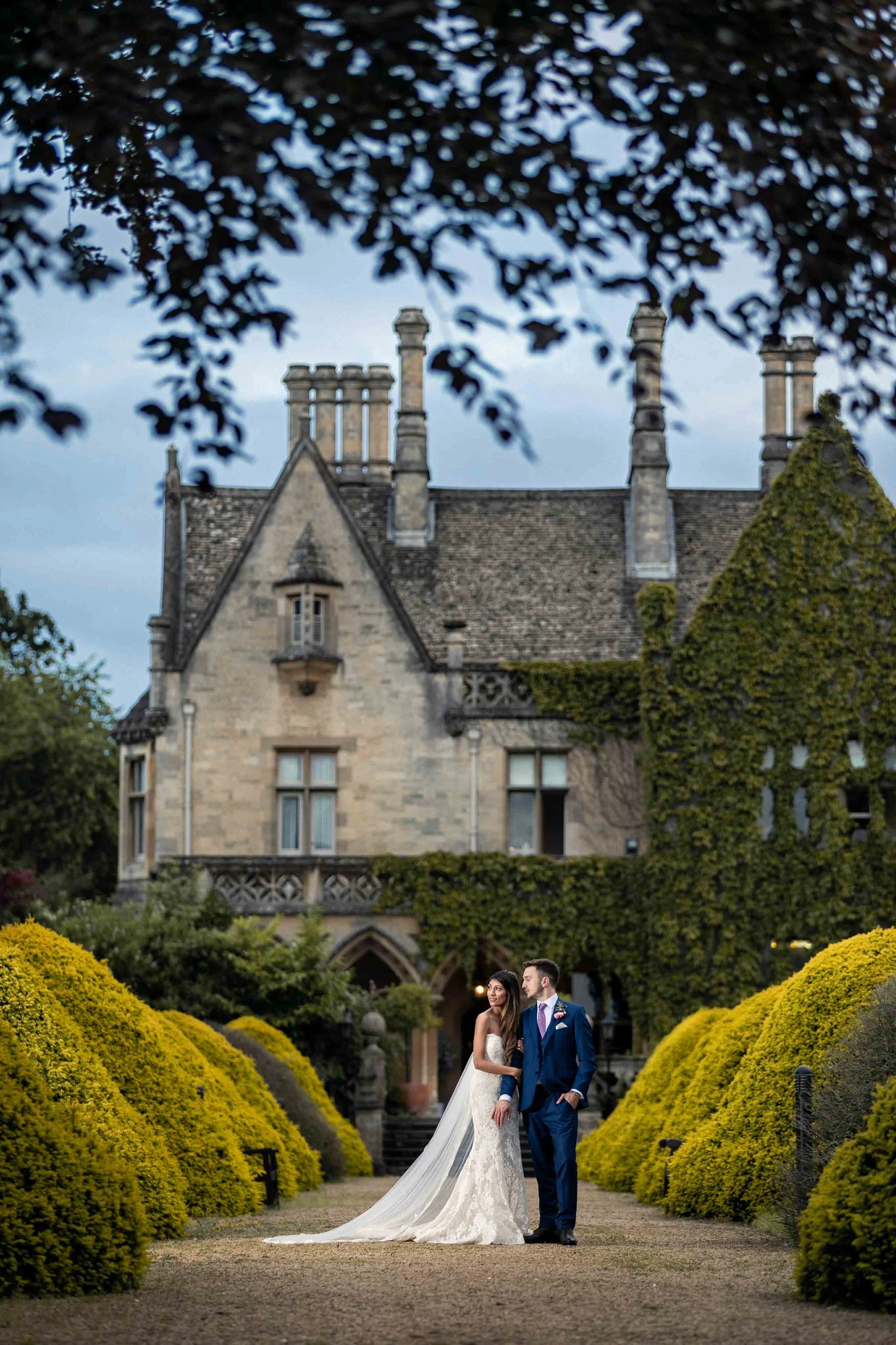 A bride and groom in wedding attire standing on a pathway in front of a large, historic castle surrounded by greenery.