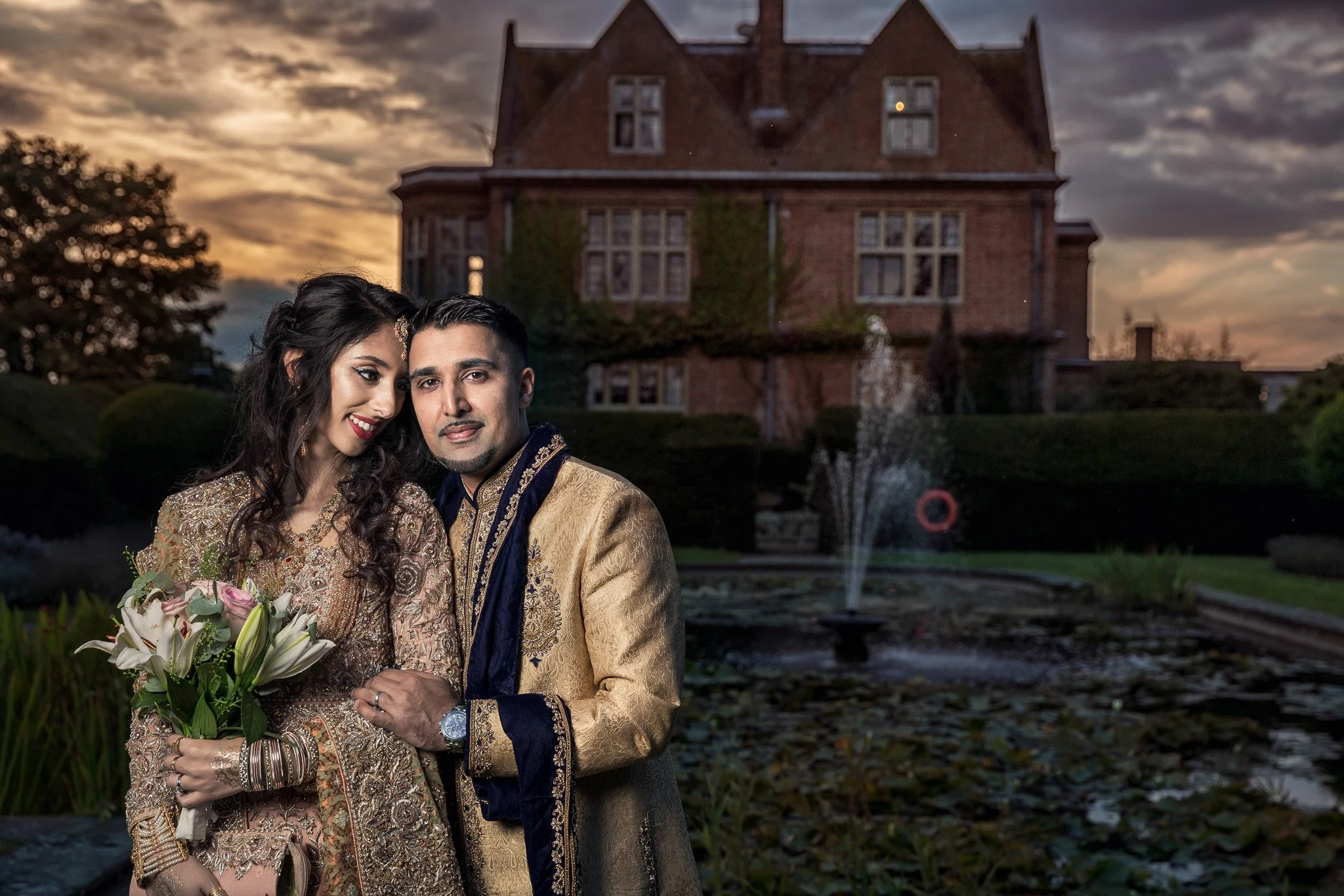 A woman and man dressed in traditional South Asian wedding attire standing close together in front of a mansion with a pond and fountain during sunset.
