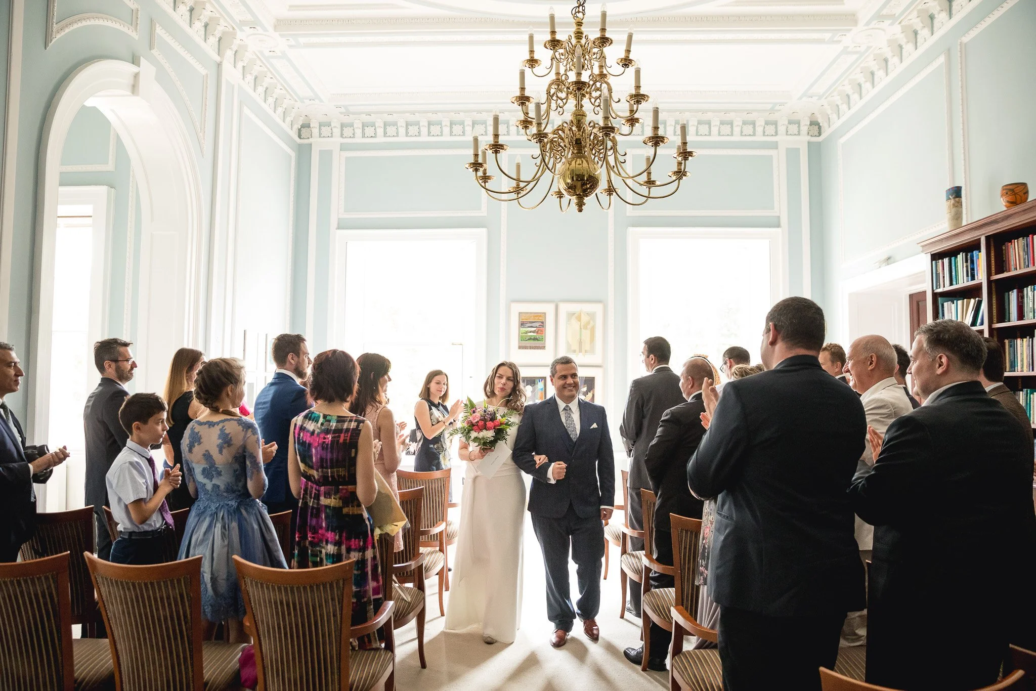 A bride and groom walking down the aisle in a wedding ceremony held in a bright, elegant room with high ceilings, large windows, and a chandelier. The bride is holding a bouquet of flowers, and the guests are standing and clapping.