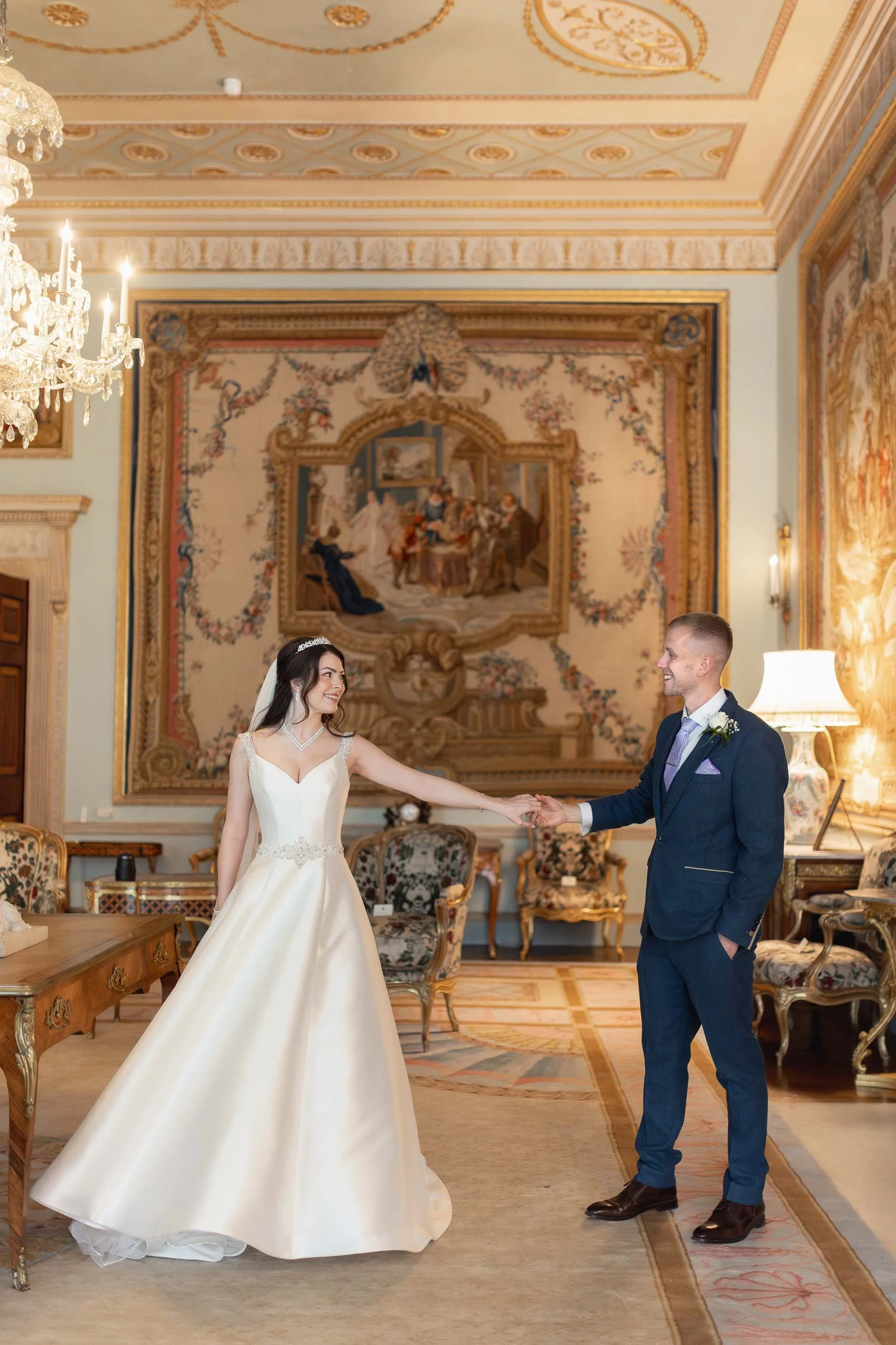 A bride and groom happily dancing in an ornate, classical-style room with luxurious decor, a chandelier, and large, decorative tapestry on the wall.