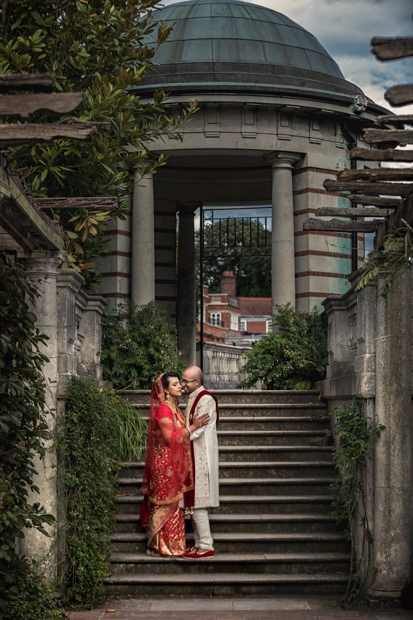 A couple dressed in traditional Indian wedding attire standing on stone steps in front of a historic building with columns and greenery.