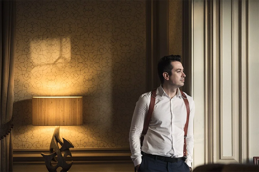 A young man with dark hair, dressed in a white shirt and suspenders, standing by a window and looking outside. The room has warm lighting from a table lamp and patterned wallpaper.