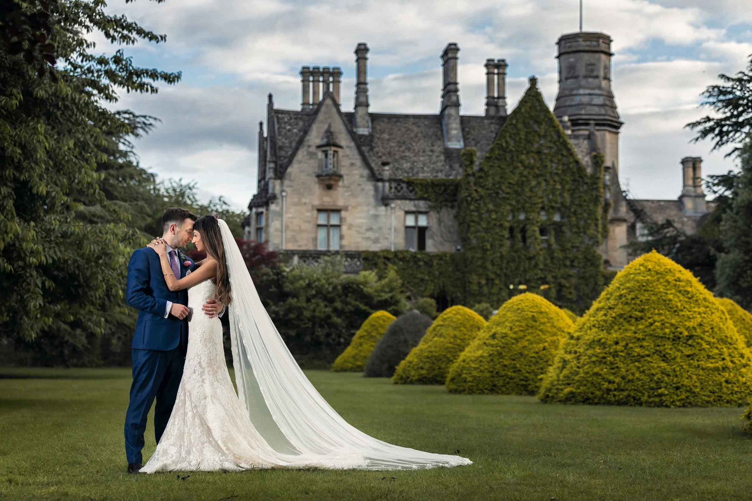 Bride in white wedding gown and long veil, groom in blue suit, standing close and holding each other on a lush green lawn in front of a Victorian-style castle with ivy-covered walls, surrounded by manicured bushes and trees.