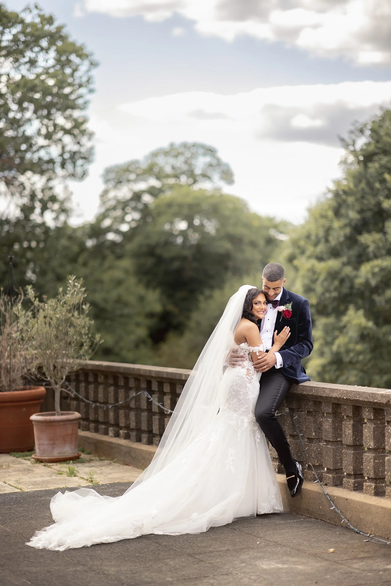 A newlywed couple in wedding attire embracing outdoors on a bridge, with trees and a cloudy sky in the background.